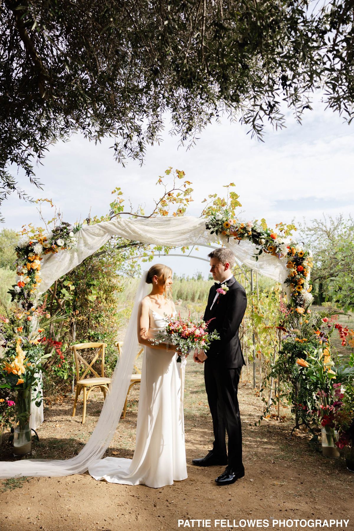 Bride and groom stand beneath floral arch with orange, red, and burgundy flowers in vineyard setting