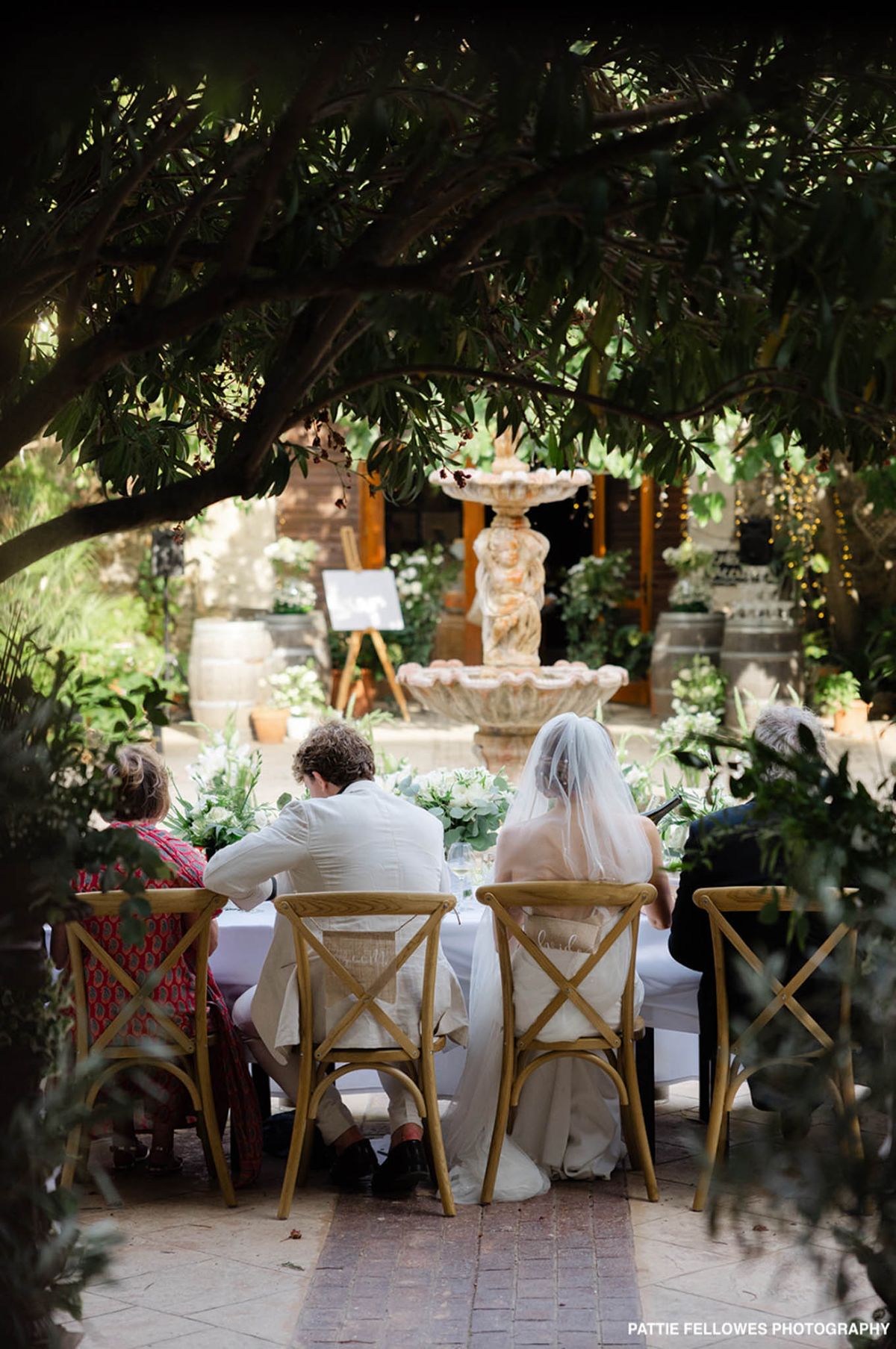 Bride and groom at sweetheart table with white florals beside stone fountain framed through tree branches
