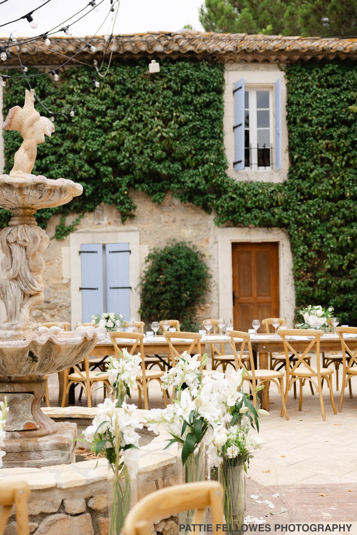 Long tables around stone fountain with white lily arrangements and string lights in Provencal mas courtyard