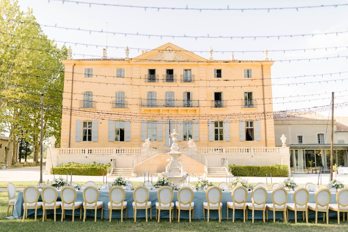 Long dusty blue table with Louis XVI chairs before ochre chateau with fountain and bistro lights overhead