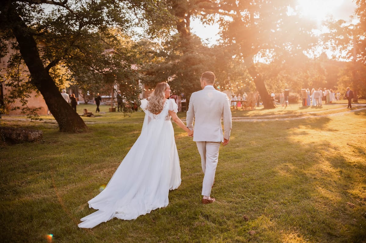 Bride and groom walking toward cocktail hour guests at golden hour with fairy-lit stone building behind