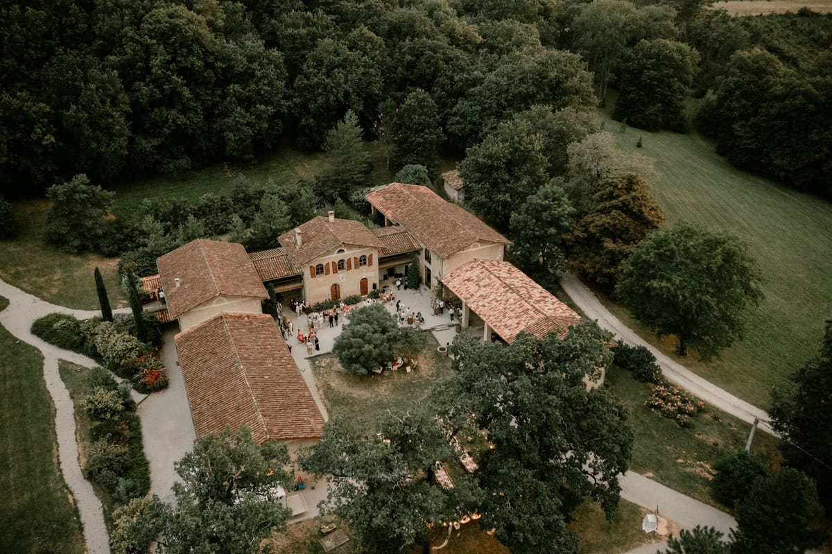 An aerial drone shot of a French country estate during what appears to be a cocktail hour or outdoor reception, with approximately 40–50 guests gathered in the central courtyard between the main stone farmhouse buildings. The property features multiple connected structures with terracotta-tiled roofs and warm ochre rendered walls, arranged around a central open-air courtyard with mature olive trees and seating areas. Guests are visible mingling across the courtyard, and string lights are partially visible in the lower portion of the image among the trees, suggesting evening lighting has been set up. The estate is surrounded by gravel driveways, manicured grounds, and dense woodland on all sides, with open fields visible to the upper right. Potential venue feature image.