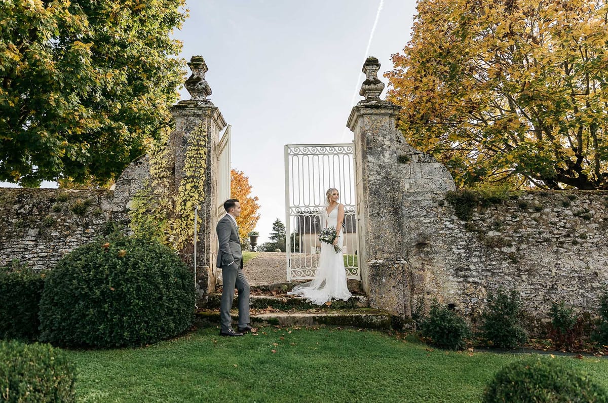 Bride in lace gown standing in wrought-iron gate facing groom on lawn at stone-walled estate