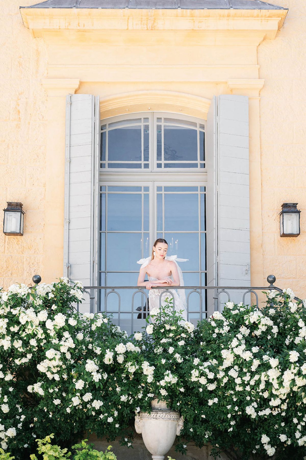 Bride in strapless lace gown with organza shoulder details on balcony of yellow chateau with roses