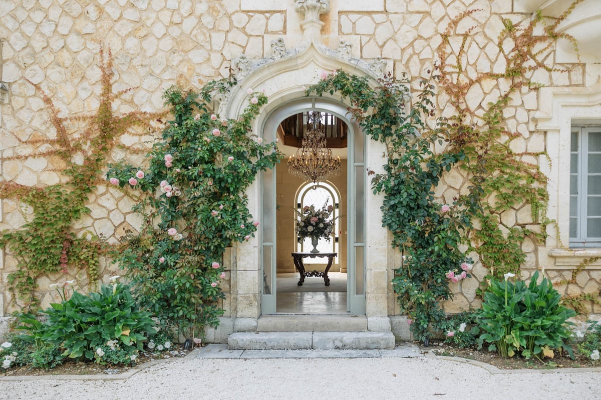 Chateau entrance with climbing roses, blue-grey doors, crystal chandelier, and blush rose arrangement in stone urn