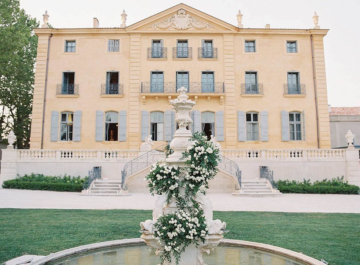 A wide exterior shot of a three-story French château with warm ochre-yellow rendered walls, grey-blue shutters, wrought iron balconies, and a classical pediment with ornate stone carvings above the roofline. In the foreground, a large stone fountain basin features a tiered central column decorated with a cascading floral installation of white roses and lush green foliage. The château's entrance is framed by a symmetrical double staircase with wrought iron railings, and small candles are placed along the base of the steps. No people are visible, suggesting this is a pre-event or venue setup shot. The decor palette is white and green with a classic, formal aesthetic. Potential venue feature image.