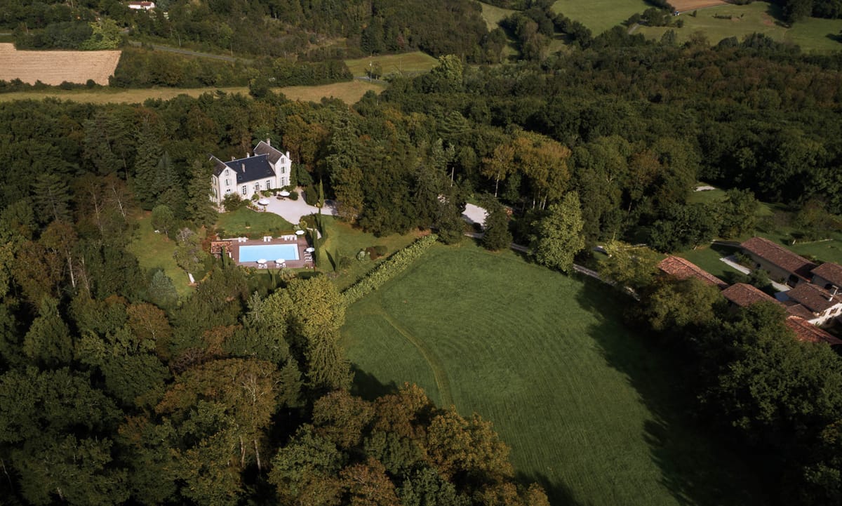 Aerial wide shot of a French château and its grounds, showing a white multi-story manor house with a dark slate roof, multiple chimneys, and classic French architectural details. The property features a rectangular outdoor swimming pool with a wooden deck surround, white sun umbrellas, and lounge furniture arranged around it. A gravel driveway with additional white umbrellas and outdoor seating is visible near the main entrance. The estate is surrounded by mature woodland on multiple sides, with an open lawn area adjacent to the pool and a large open field stretching to the right of the property. Secondary outbuildings with terracotta tile roofs are visible to the lower right. No people are visible in the image. Potential venue feature image.