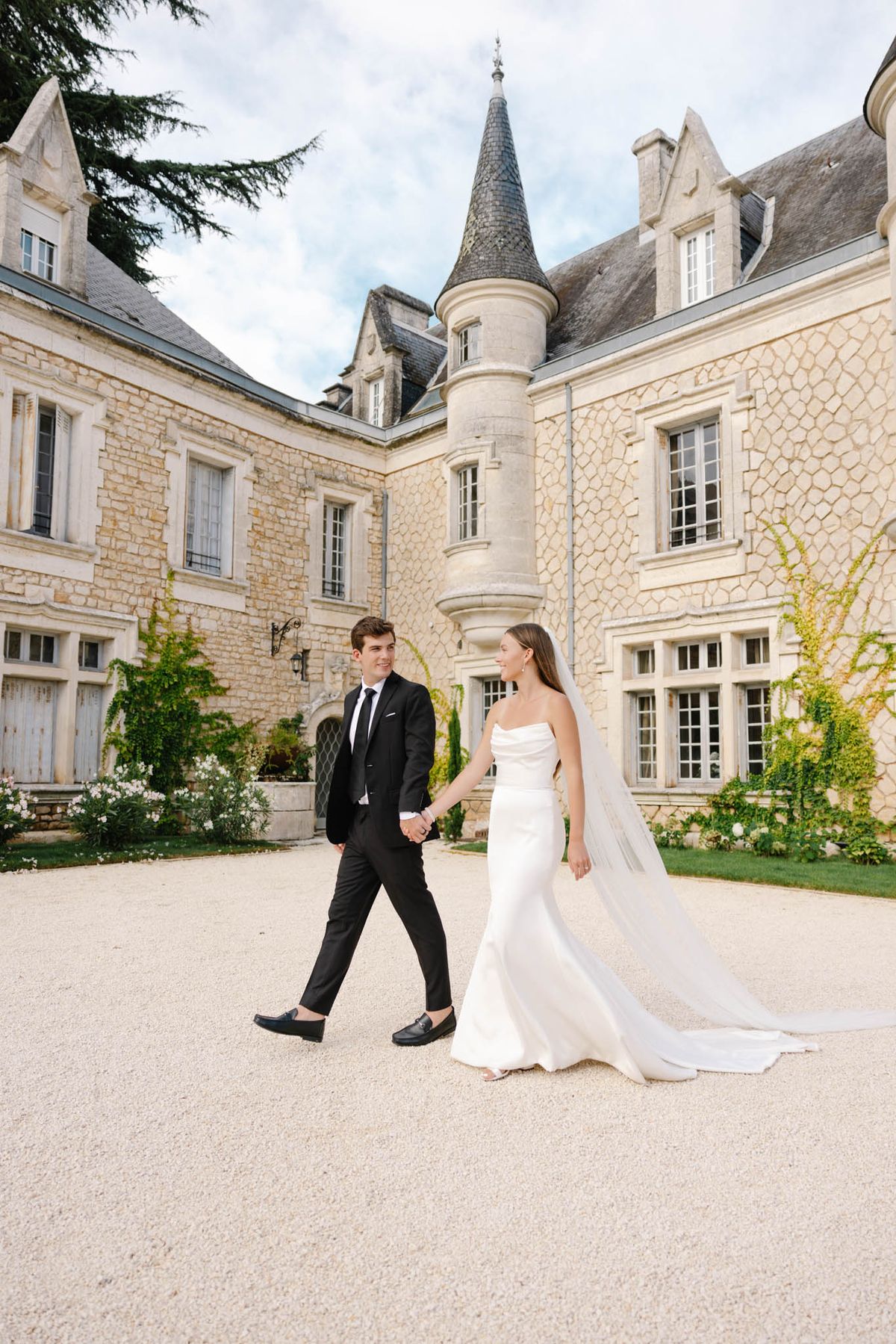 A couple portrait taken outdoors on the gravel courtyard of a French château featuring a distinctive conical turret and cream limestone facade with climbing greenery. The bride wears a strapless, fitted ivory satin gown with a long cathedral-length veil trailing behind her, while the groom is dressed in a slim black suit with a black tie and black loafers. The two are walking hand-in-hand across the courtyard, with the bride glancing toward the groom and smiling. The overall styling is clean and modern against the classic French château architecture. Wide portrait shot. Potential venue feature image.