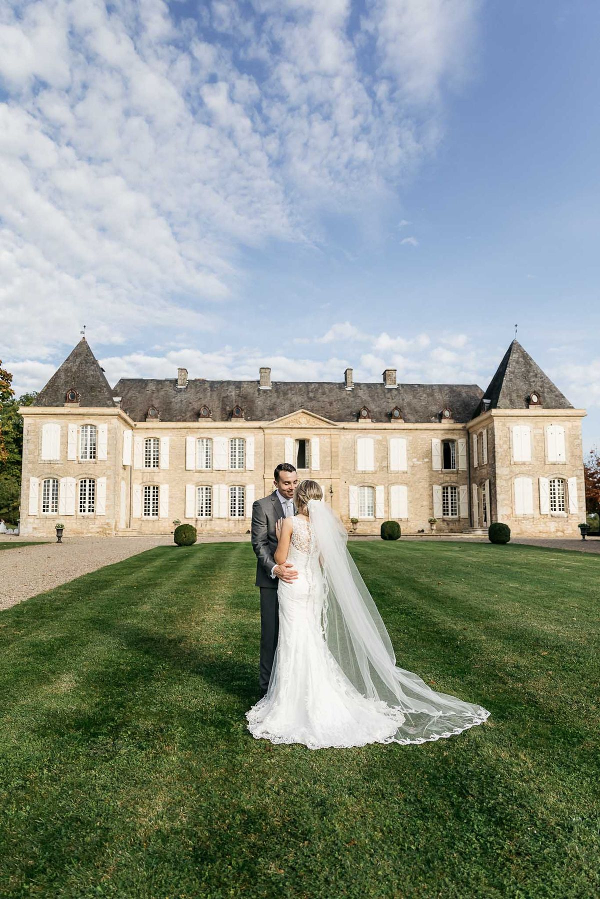 Couple on lawn with lace-trimmed cathedral veil before symmetrical cream stone chateau with slate turrets