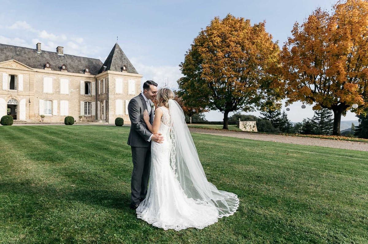 A couple portrait taken outdoors on the grounds of a French château during autumn. The bride wears a fitted ivory lace gown with a deep V-back and a cathedral-length veil with a lace-trimmed edge that fans out across the lawn; the groom wears a charcoal grey suit with a light tie. The two are facing each other in an embrace, smiling, positioned on a well-maintained lawn with the limestone château facade and its dark slate mansard roof visible in the background. The composition is a medium-wide shot taken from slightly behind and to the side of the couple, with the château anchoring the left side of the frame and a large tree with deep amber and orange autumn foliage filling the right side.