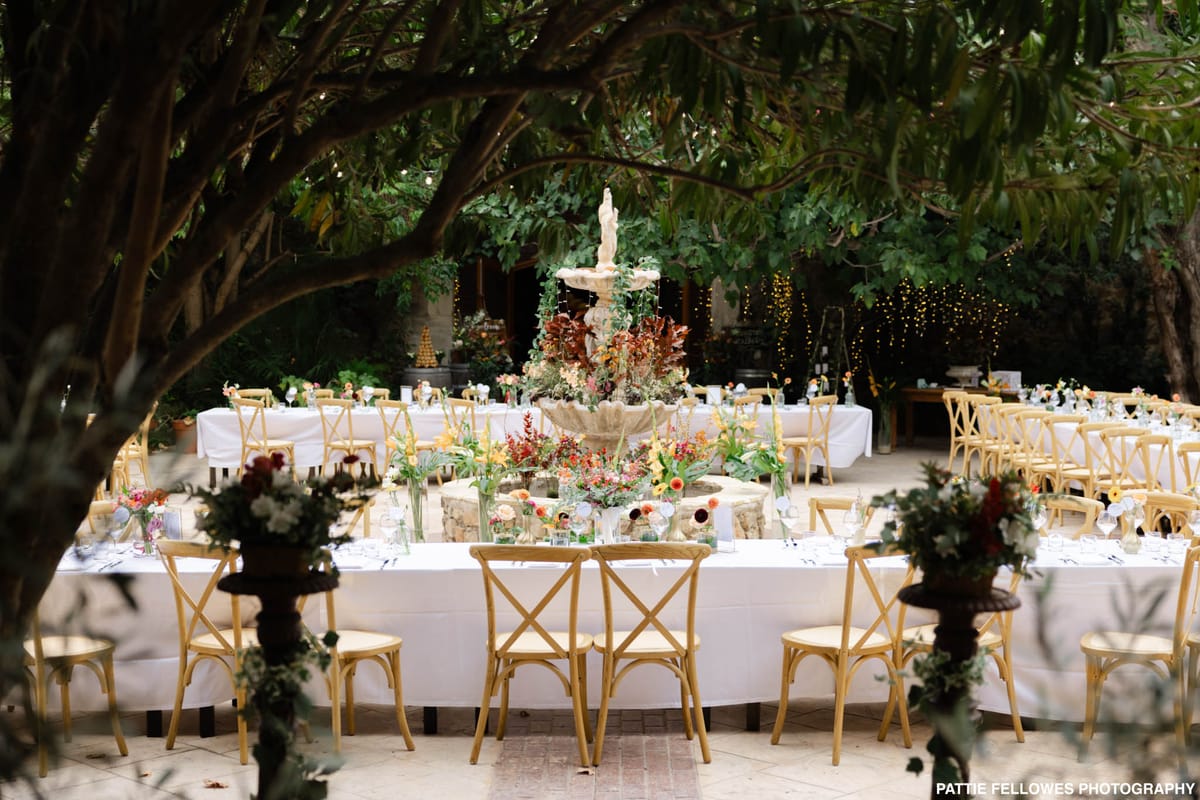 An outdoor wedding reception setup in a courtyard centered around a large stone tiered fountain decorated with trailing greenery and burgundy foliage. Long rectangular tables with white linen cloths are arranged around the fountain, seated with natural wood cross-back chairs. The floral arrangements are abundant and colorful, featuring orange, yellow, red, coral, and burgundy blooms — including what appear to be ranunculus, snapdragons, and tropical-style stems — displayed in clusters of glass bud vases along the table runners. Additional potted arrangements with white flowers and greenery are placed at the ends of the tables. Warm fairy lights are strung through the trees in the background, and a tiered cake on a stand is visible to the rear left. The overall decor palette is warm and saturated with a garden-inspired, bohemian style. Wide establishing shot capturing the full reception layout. Potential venue feature image.