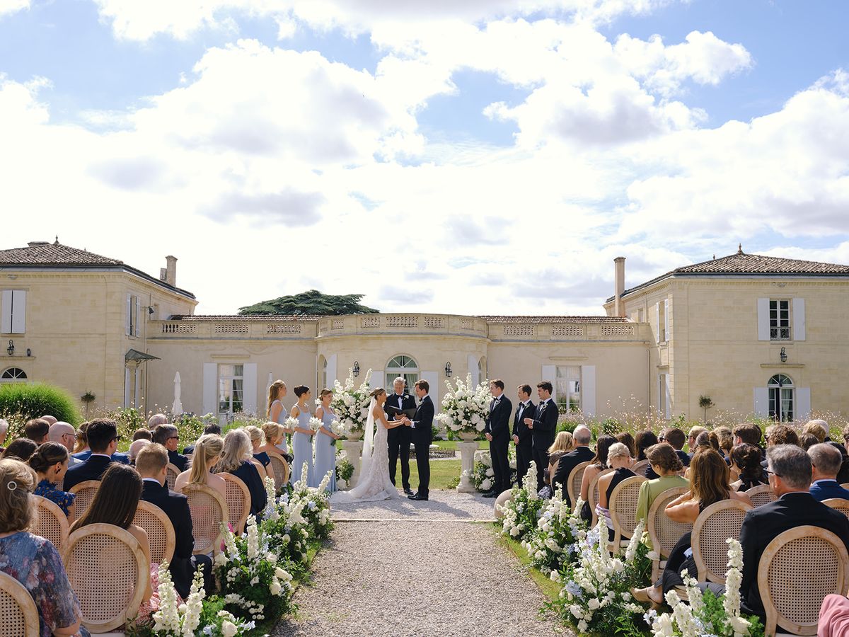 Wide elevated shot of outdoor ceremony in château courtyard with 80-100 guests and white floral aisle arrangements