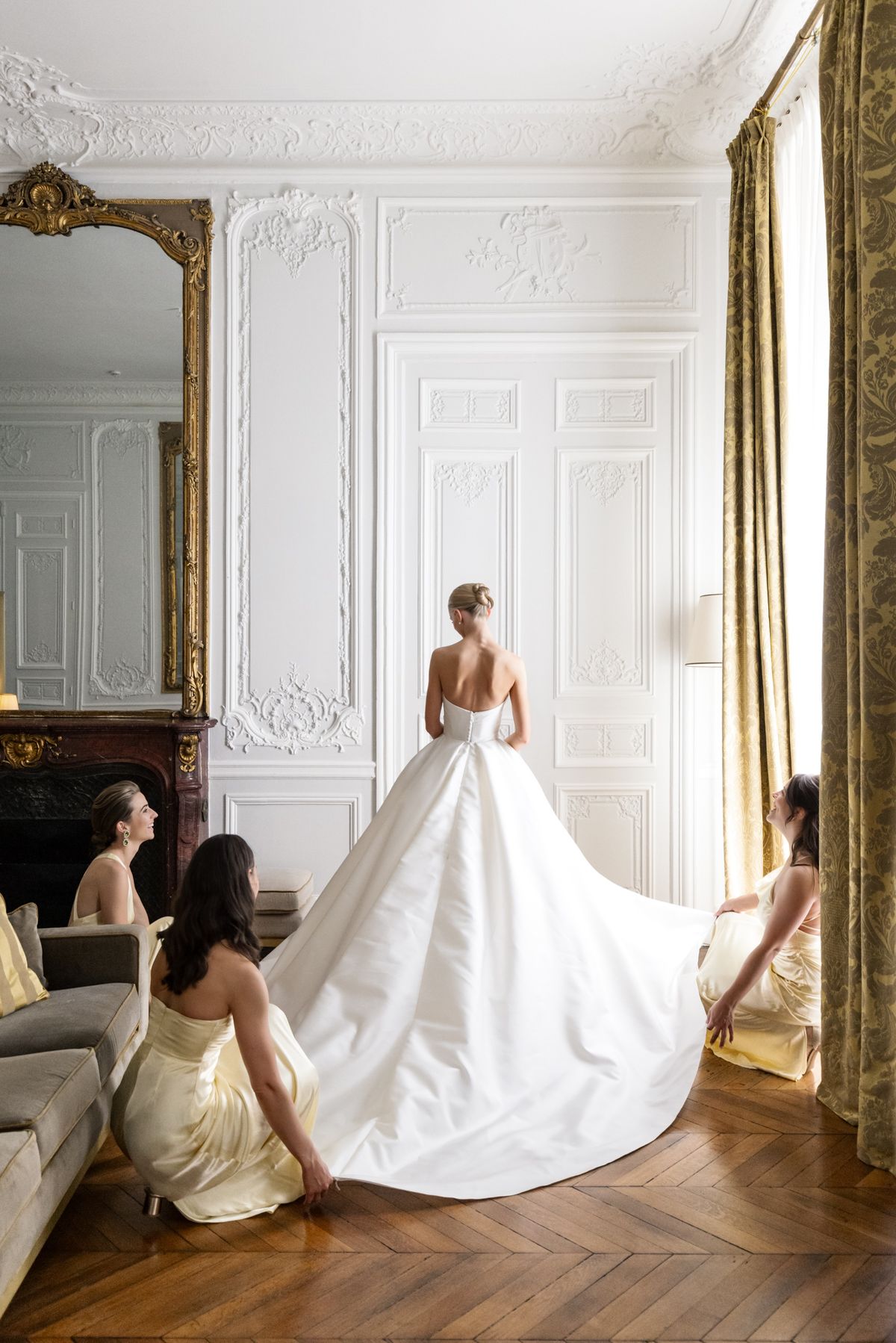 Bride in ivory ball gown from behind in Parisian salon with panelled walls, gold mirror, and two bridesmaids in pale yellow