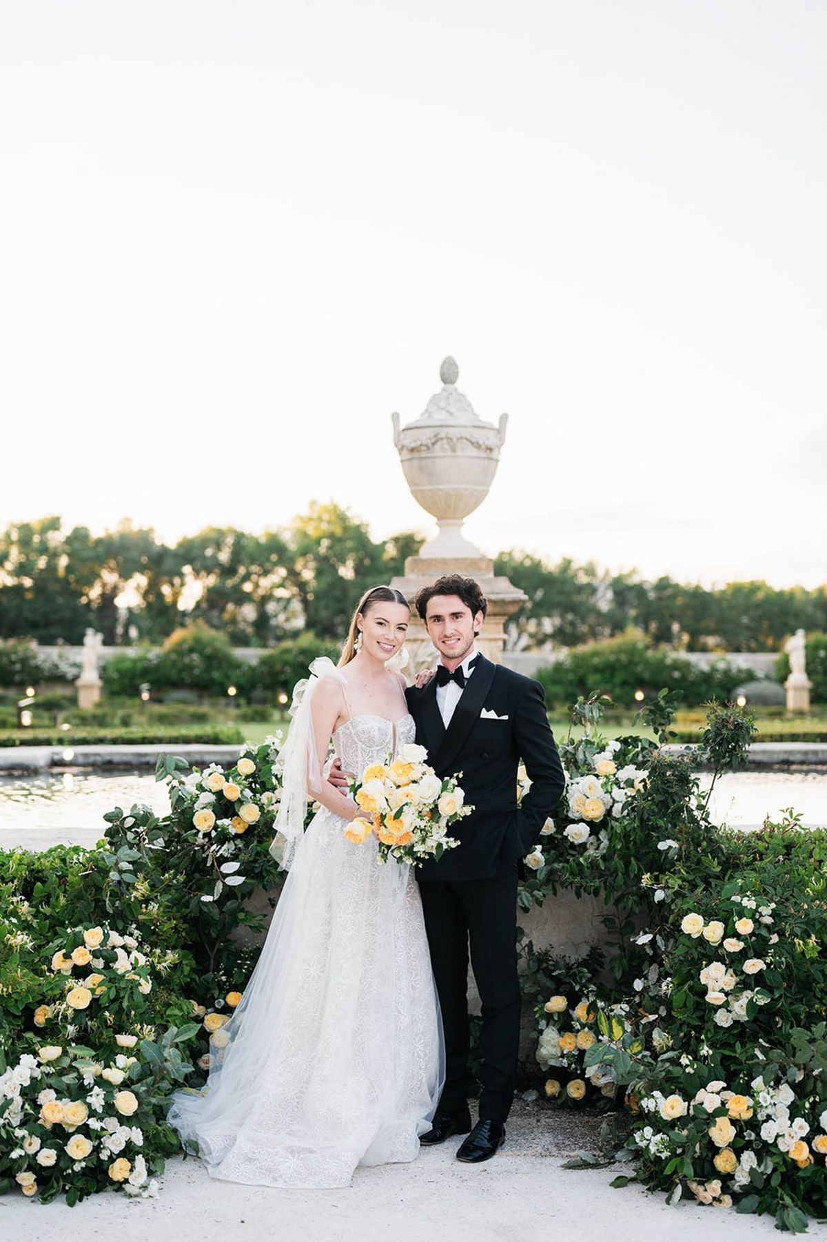 A couple poses together for a portrait in a formal French garden at dusk, standing in front of a large stone ornamental urn on a pedestal beside a reflective pool. The bride wears a fitted lace A-line gown with a sweetheart neckline, thin straps, a full tulle skirt, and a long veil, and holds a loose bouquet of yellow, peach, and white roses with greenery. The groom is dressed in a black double-breasted tuxedo with a black bow tie and white pocket square. They are framed by large floral ground arrangements of yellow and white roses with lush green foliage that line a low stone balustrade. The overall floral palette is yellow, peach, and ivory, consistent with a classic formal garden wedding style. This is a mid-length portrait shot.