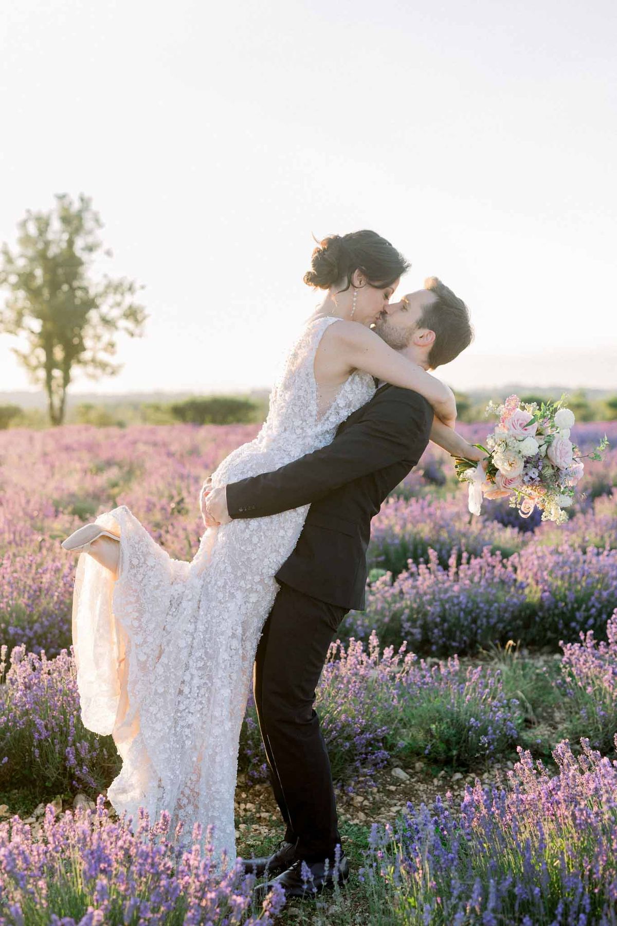 Groom lifting and kissing bride in lavender field at golden hour, bride in ivory lace gown holding blush and cream bouquet