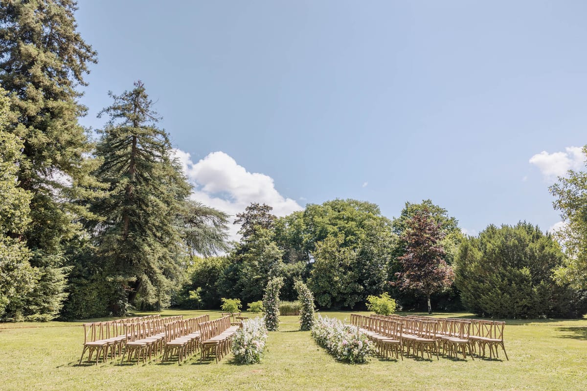 An outdoor ceremony setup photographed in a wide shot on a large lawn surrounded by mature trees. Rows of natural wood cross-back chairs are arranged in two sections flanking a central aisle, accommodating approximately 80–100 guests. The aisle is lined with low floral arrangements featuring white and soft pink blooms, and a circular floral arch stands at the altar end, decorated with white and blush flowers and greenery. The overall styling is garden-romantic with a natural, airy palette of whites, blush, and greens. No people are present — the space is set up and awaiting the ceremony. Potential venue feature image.