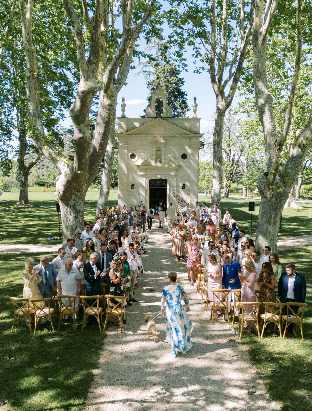 An outdoor wedding ceremony is underway in the grounds of a French estate, with guests seated in natural wood cross-back chairs arranged in two rows along a gravel aisle. A small stone chapel with classical architectural detailing — including a pediment, decorative urns, and a bell niche — serves as the ceremony backdrop, flanked by large white floral arrangements. Approximately 80–100 guests are seated and standing, dressed in summer attire including a Scottish tartan kilt, a cobalt blue suit, and various pastel and printed dresses. In the foreground, a woman in a white and blue floral floor-length dress walks a small dog up the aisle toward the altar. The shot is taken from an elevated wide angle, capturing the full symmetry of the aisle and ceremony setup. Potential venue feature image.
