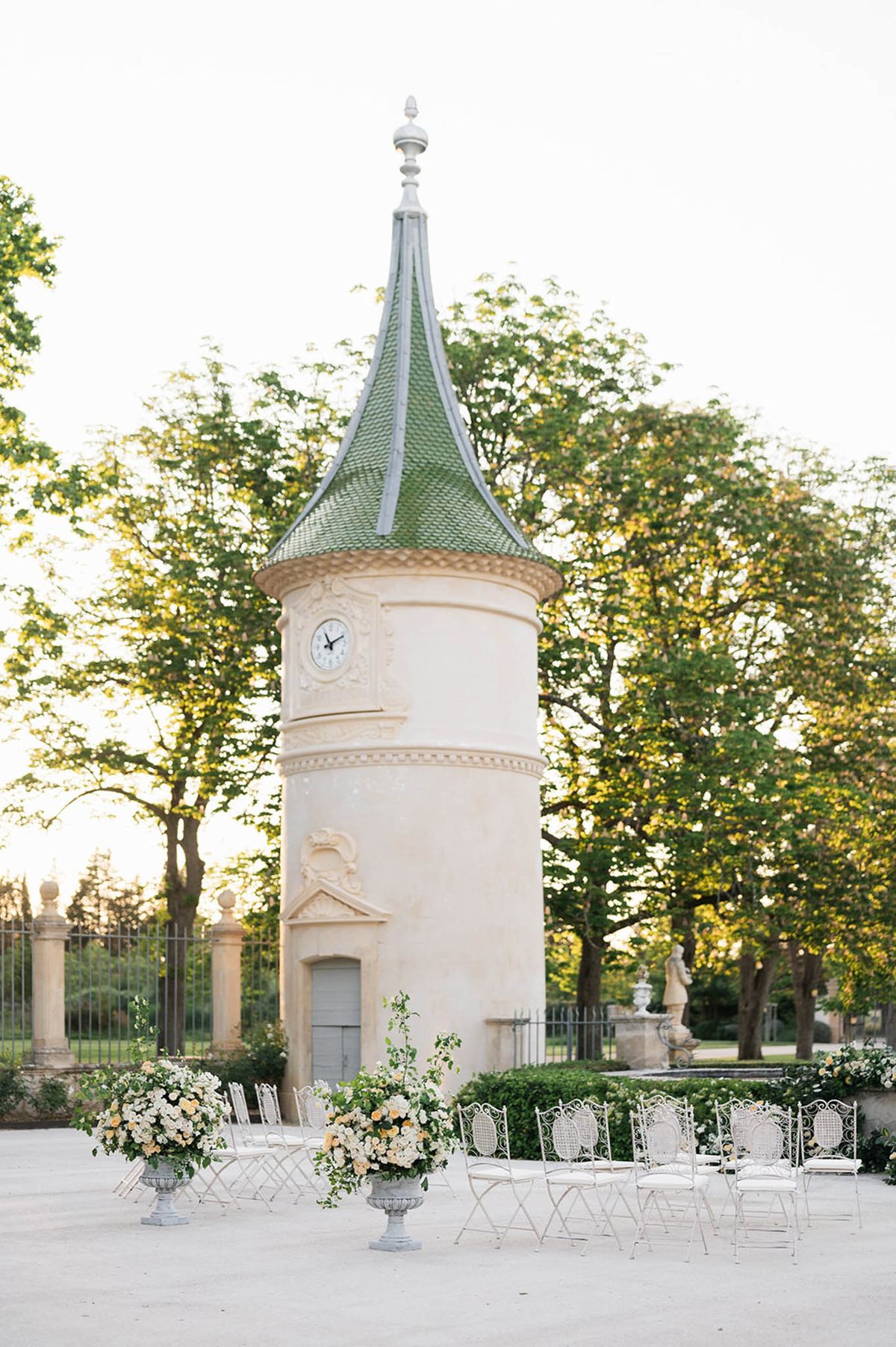 White wrought-iron chairs and rose-filled stone urns before round chateau tower with clock and green roof