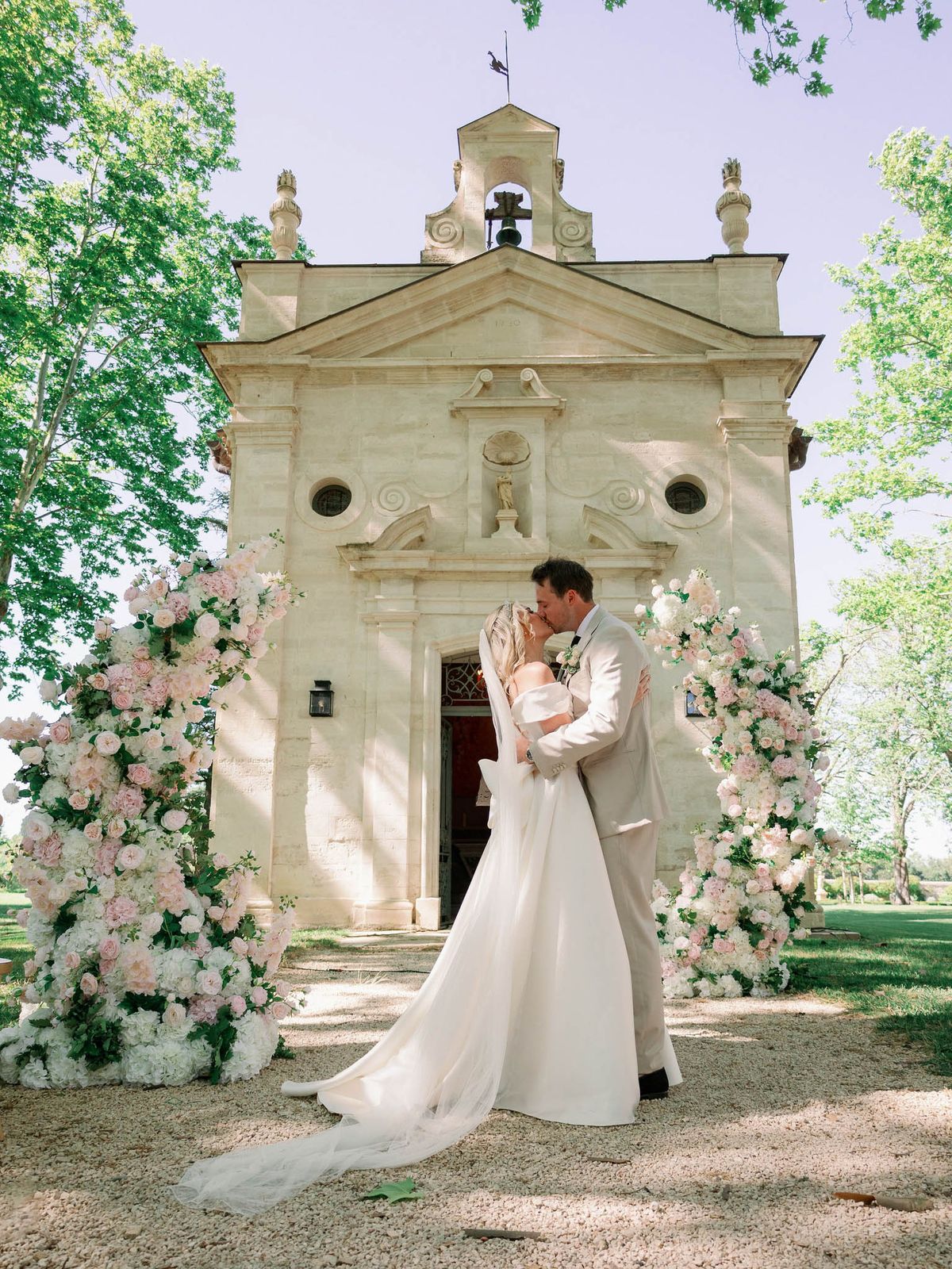 A bride and groom share a kiss outdoors in front of a small French Baroque-style chapel with a stone façade, bell tower, and classical pediment detailing. The couple is framed by two tall floral columns flanking the chapel entrance, composed of blush pink garden roses, white hydrangeas, and soft greenery. The bride wears an off-the-shoulder white gown with a cathedral-length train and veil, while the groom wears a light beige suit with a white floral boutonnière. The shot is a wide portrait taken on a gravel path, capturing the full chapel facade and the couple in an embrace at its center. Potential venue feature image.