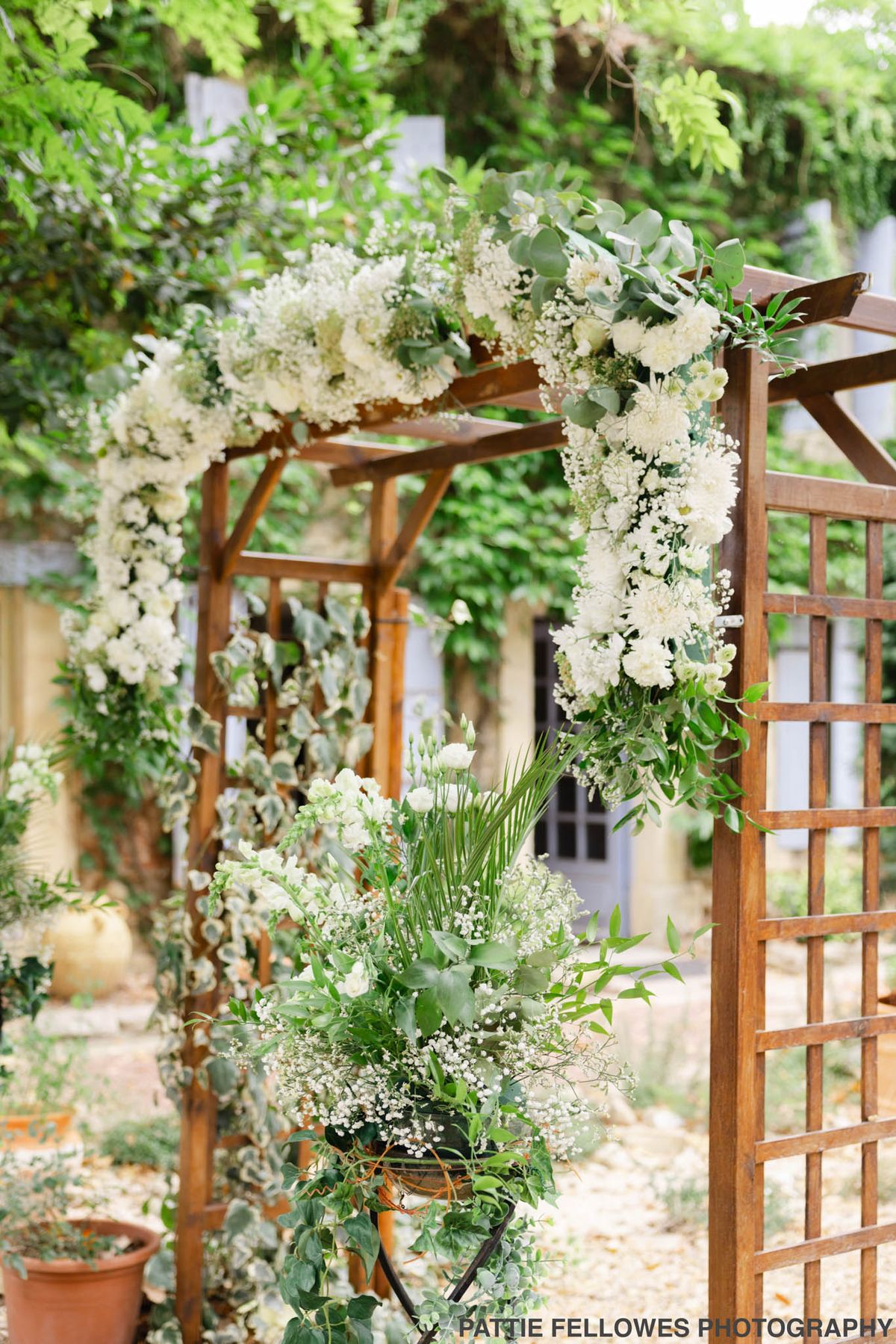 A close-up detail shot of an outdoor wedding ceremony arch decorated in a green and white floral palette. The wooden pergola-style arch is adorned with garlands of white chrysanthemums, baby's breath, white lisianthus, and eucalyptus foliage draped along the uprights and across the top beam. At the base of one post, a large floral arrangement in a dark urn features additional white blooms, baby's breath, palm fronds, and trailing greenery including ivy. The overall styling is garden-romantic with a natural, lush aesthetic. No people are visible in the frame.