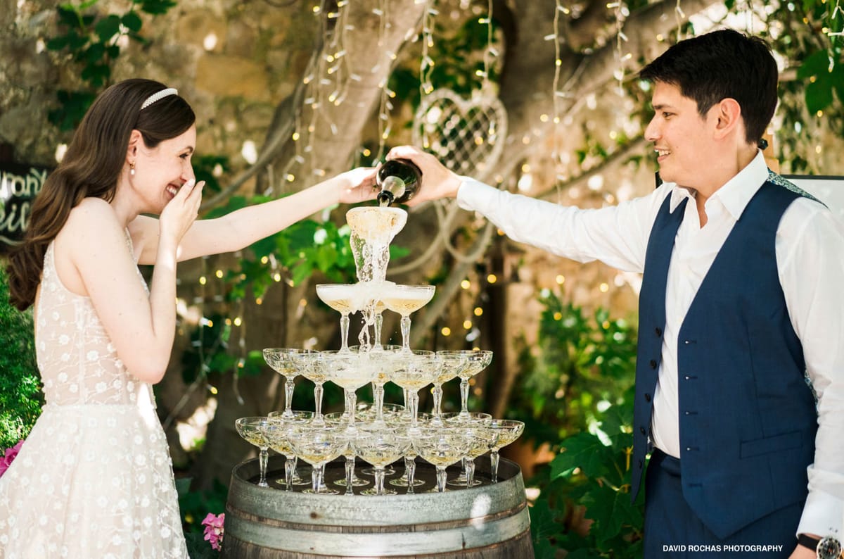 The bride and groom are pouring champagne into a coupe glass tower during an outdoor reception, set on top of a wooden wine barrel. The bride wears a sleeveless white gown with floral embroidery detail and a thin pearl headband, and reacts with laughter as champagne splashes from the top of the tower. The groom wears a white dress shirt with a navy blue vest and dark trousers. The background features a stone wall covered in greenery and warm fairy lights draped through tree branches, creating a garden setting with a rustic French countryside aesthetic. The shot is a mid-range two-person portrait capturing the action and the couple's candid reactions.