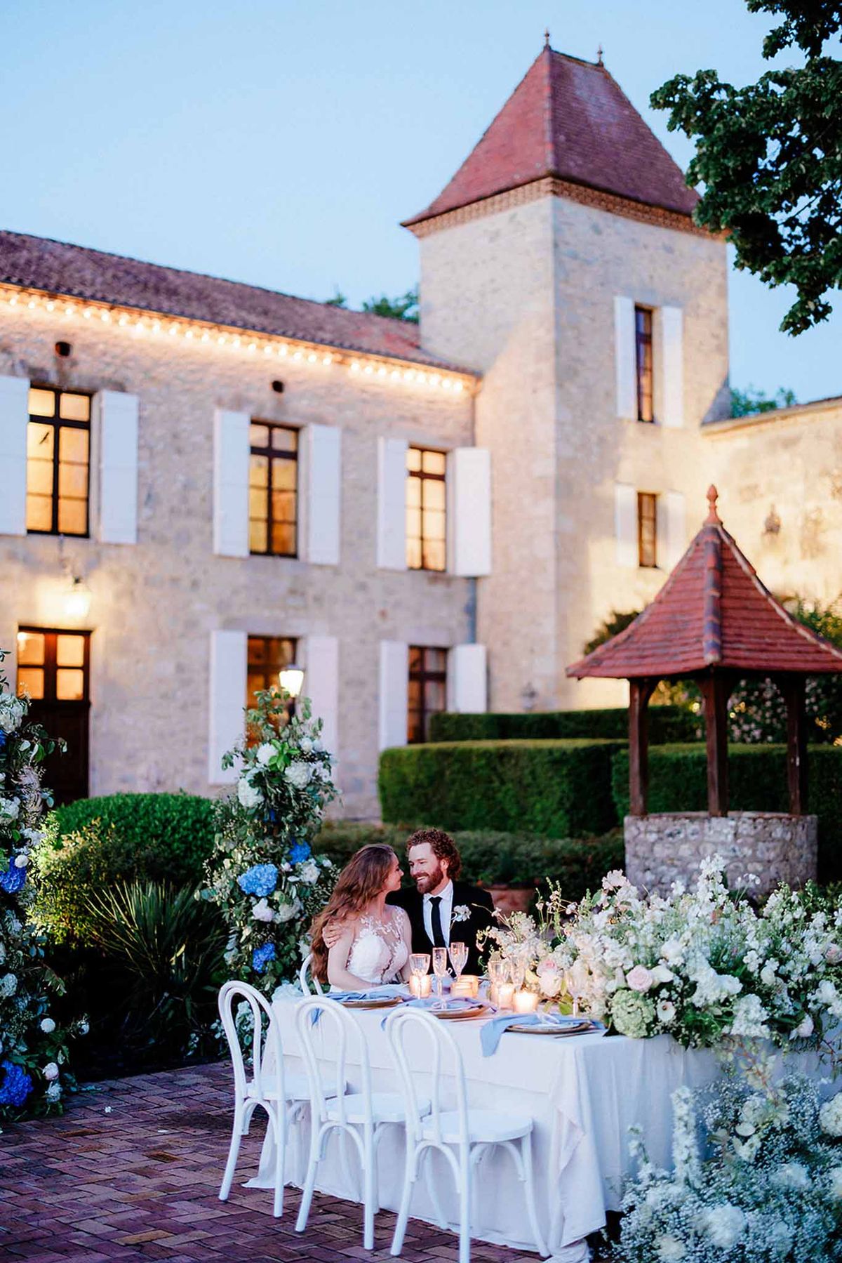 Intimate reception table for two at dusk in château courtyard with blue delphiniums and lit architectural backdrop