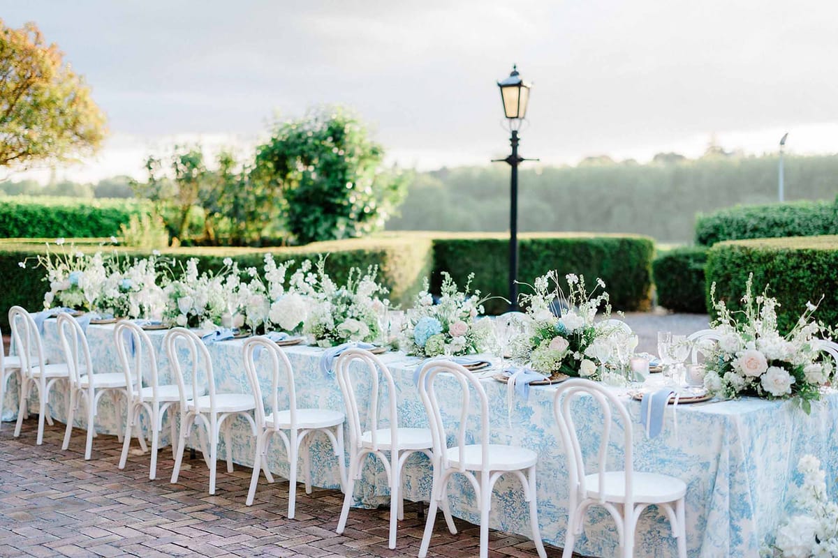 Long outdoor reception table with pale blue linens, white rose centerpieces, and bentwood chairs