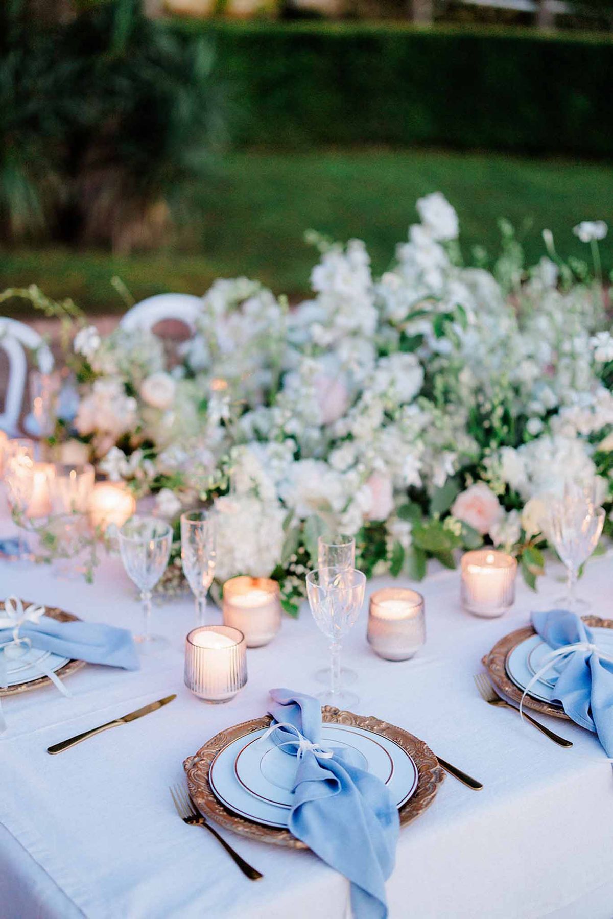 Outdoor reception table with gold charger plates, blue linen napkins, and lush blush and white floral garden backdrop