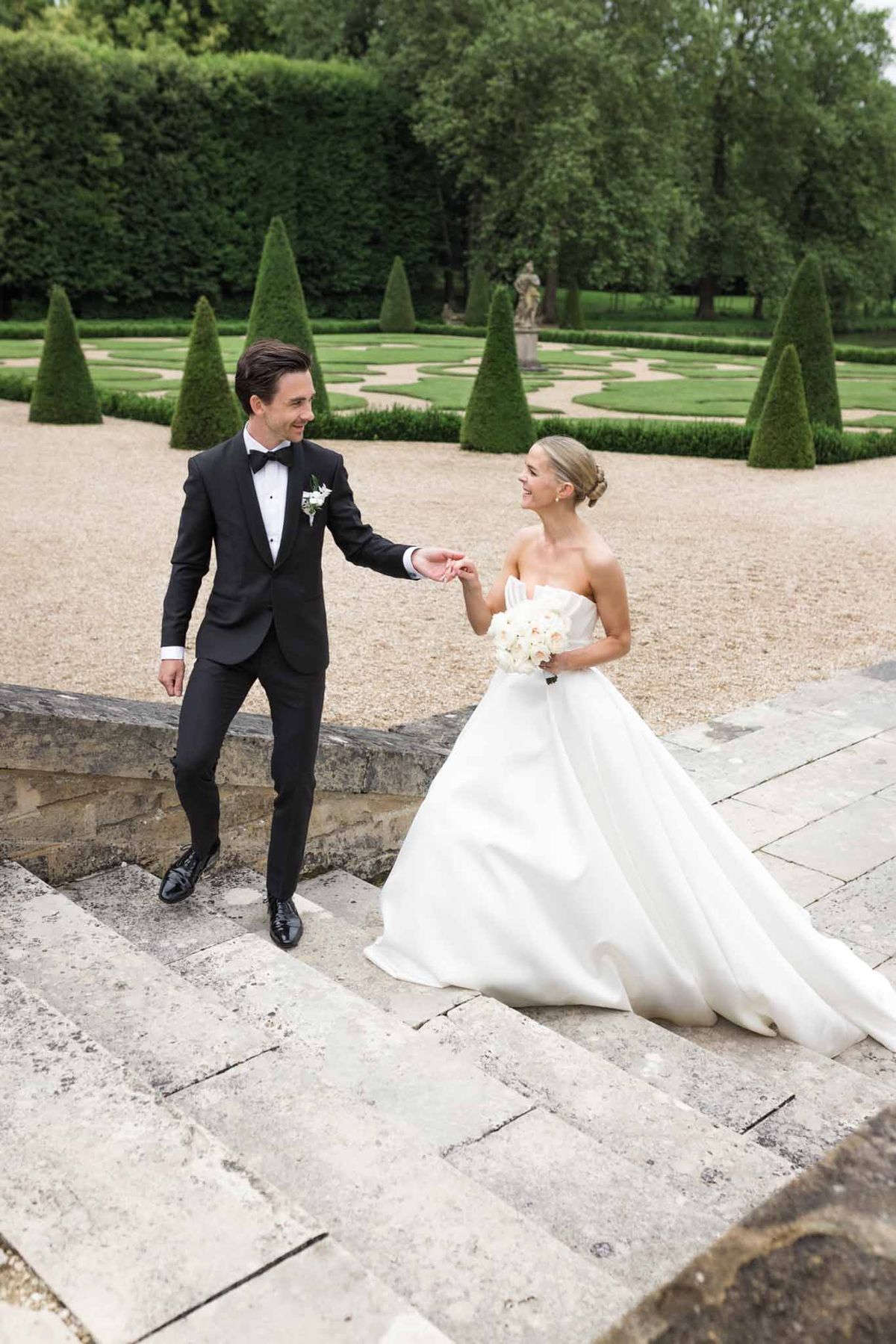 Bride in white ball gown and groom in black tuxedo walk gravel path with topiary cones and garden statue