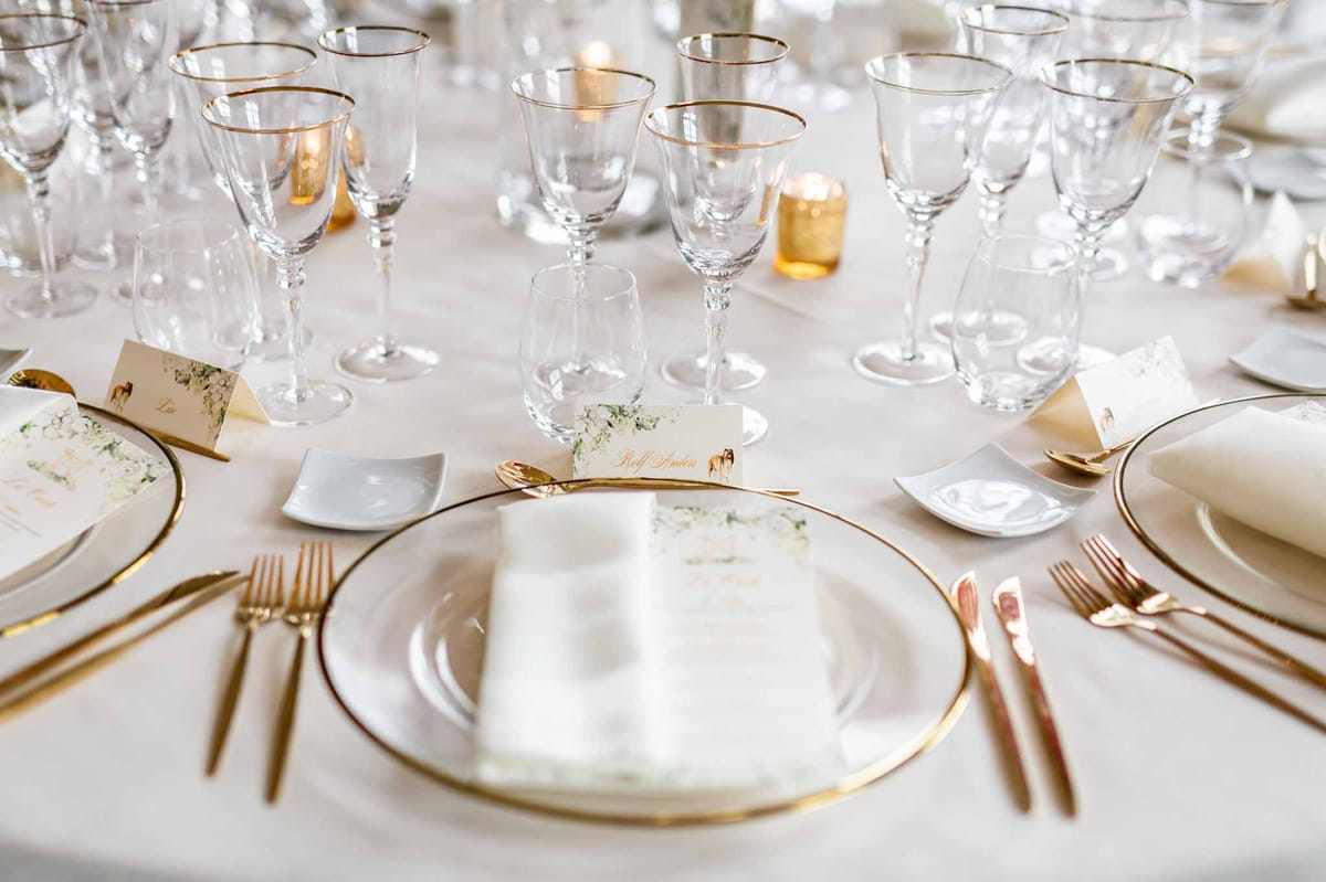 Close-up of reception table setting with gold-rimmed chargers, gold flatware, crystal glasses, and white place cards