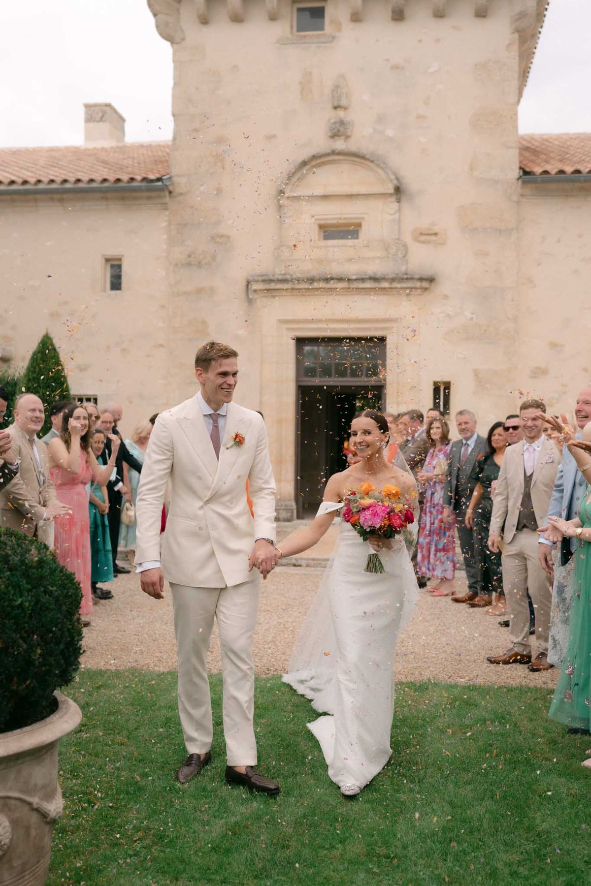 A confetti exit shot taken outdoors in front of a French château with limestone stone architecture, a decorative arched doorway, and terracotta roof tiles. The couple walks hand-in-hand through a corridor of approximately 30 guests who are throwing multicolored confetti in shades of orange, pink, and yellow. The groom wears a cream double-breasted suit with a dusty pink tie and an orange and pink boutonnière; the bride wears an off-the-shoulder fitted white gown with a short train and carries a bold bridal bouquet of hot pink, orange, and coral blooms including what appear to be dahlias and ranunculus. Guests are dressed in a colorful mix of formal attire including teal, pink, and floral dresses. The composition is a mid-length portrait shot centered on the couple with the château façade as the backdrop. Potential venue feature image.