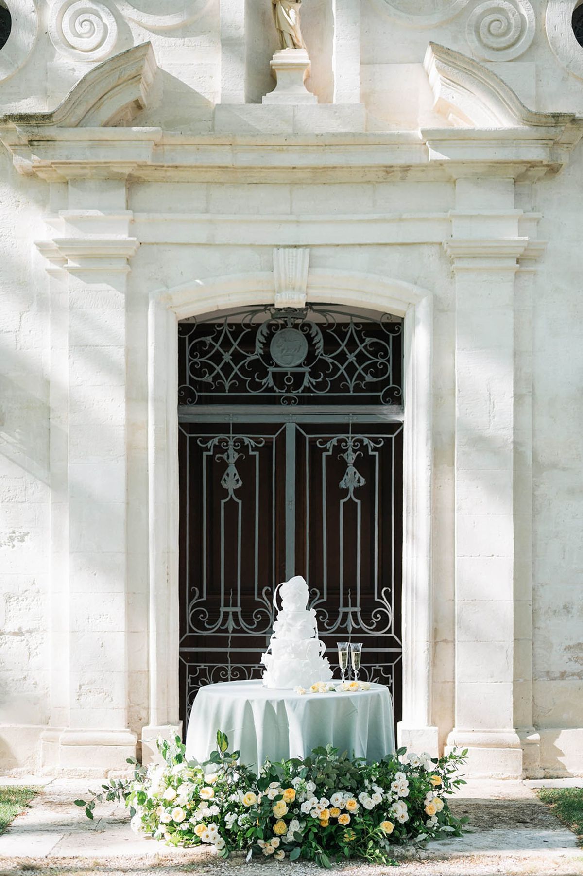 White ruffled wedding cake with champagne flutes and floral arrangement before ornate chateau stone doorway