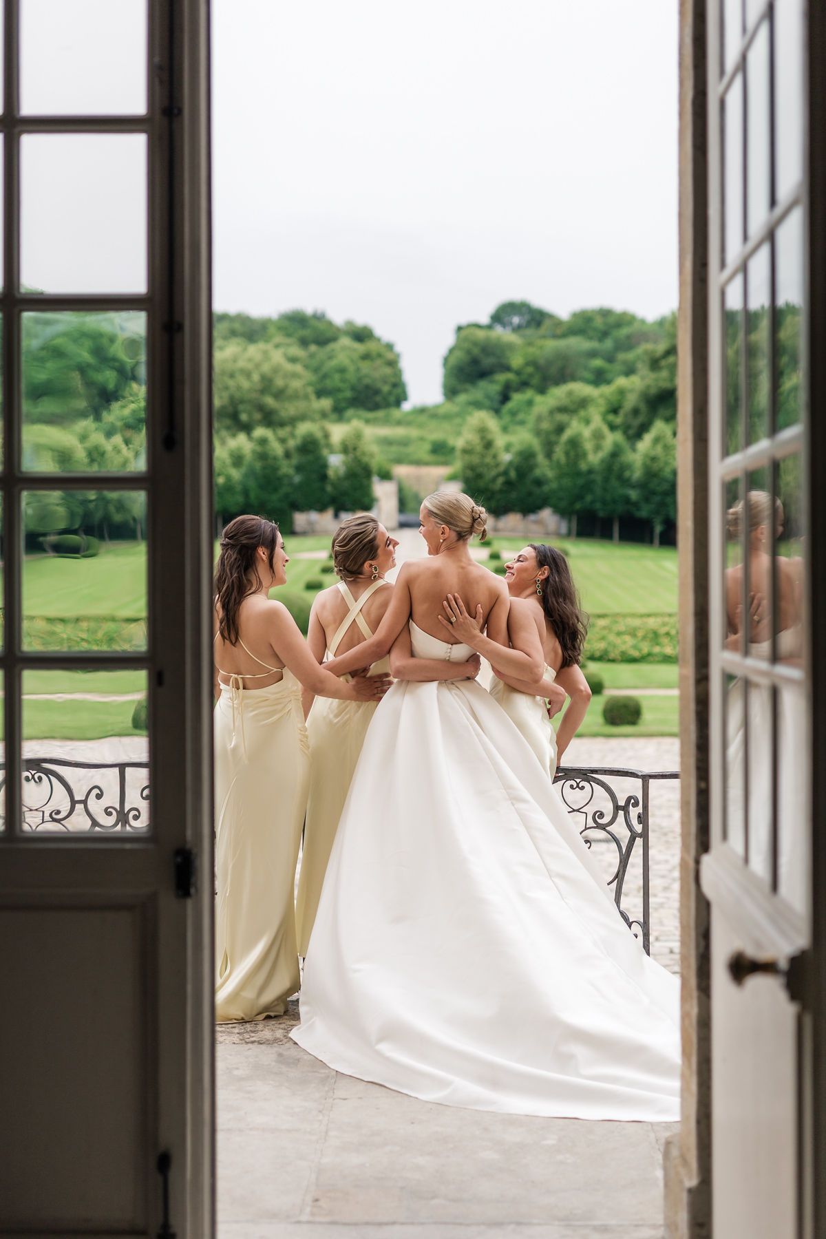 Bride in ivory backless gown and bridesmaids in champagne slip dresses viewed from behind through French doors