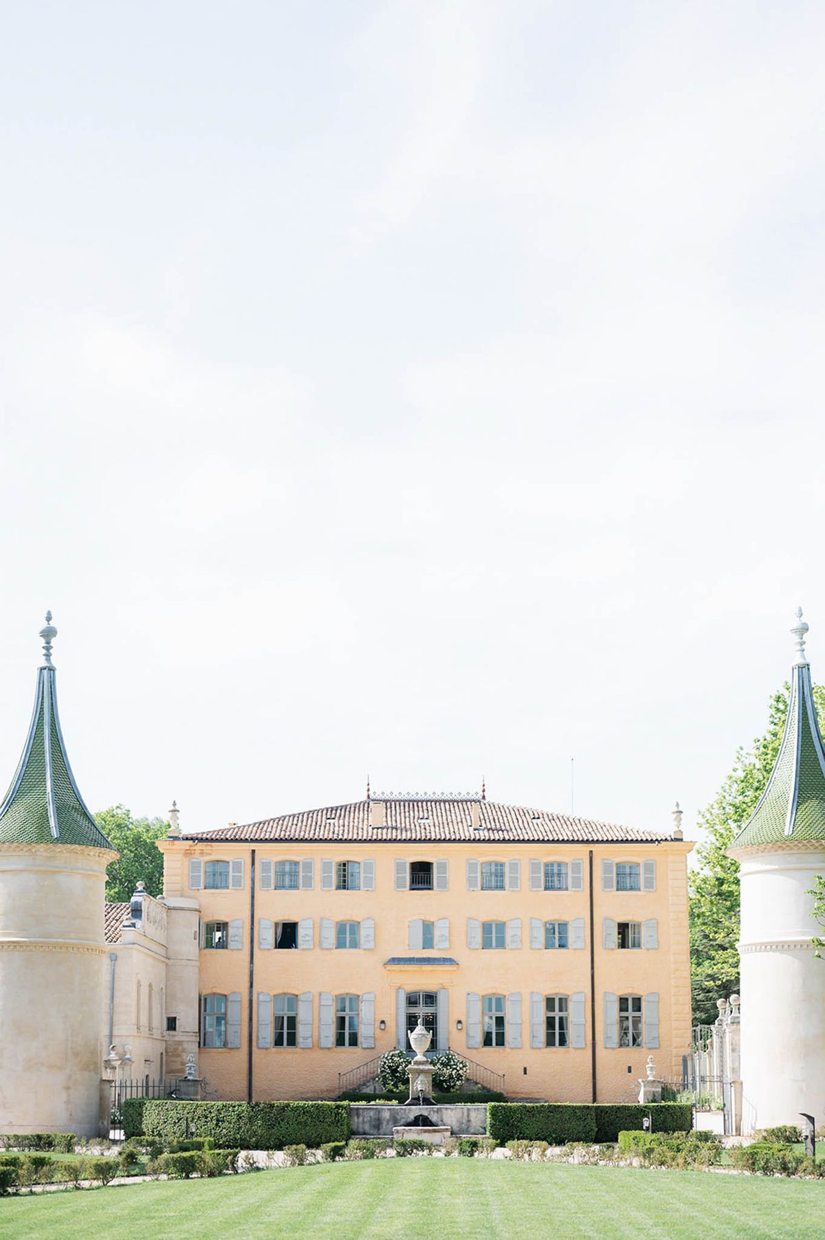 Wide exterior shot of a French château with warm ochre-yellow rendered façade, pale blue-grey shuttered windows across three stories, and a terracotta tile roof with decorative iron cresting. Two white cylindrical gatehouse towers with distinctive green pointed roofs flank the foreground, framing the main building symmetrically. A stone urn fountain sits at the base of the château's central entrance staircase, flanked by trimmed boxwood hedging and formal parterre gardens. No people are visible in the image. Potential venue feature image.