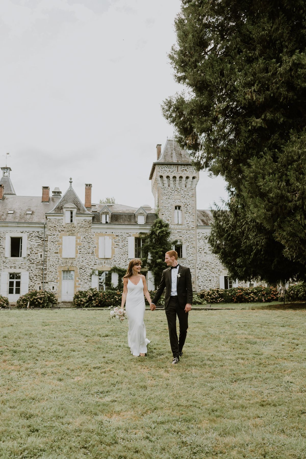 Bride and groom walking hand in hand across lawn smiling at each other with stone chateau and tower behind