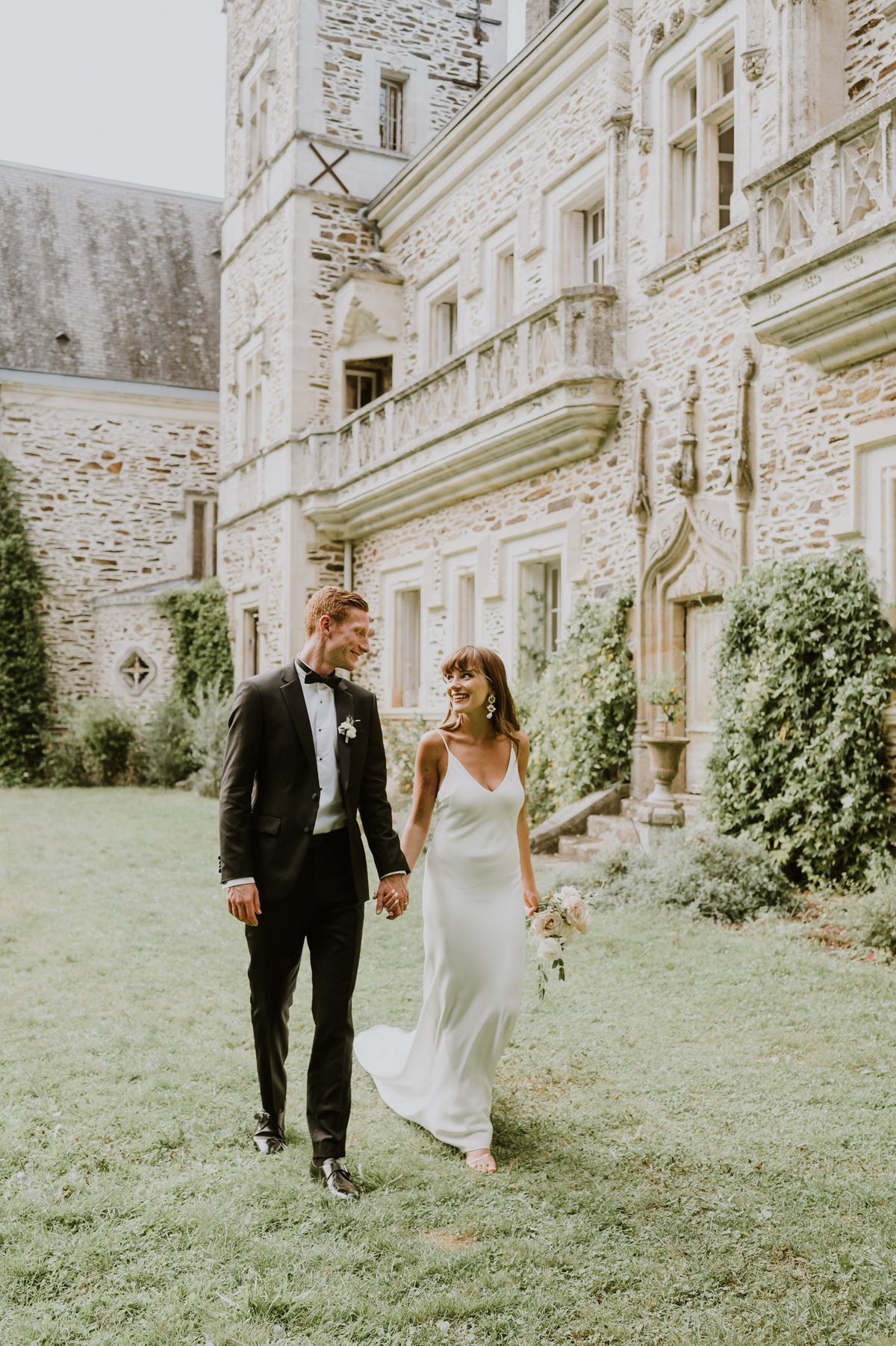 A couple portrait taken outdoors on the grounds of a French château, with the stone façade of the multi-story castle building visible in the background. The bride wears an ivory satin slip-style gown with thin spaghetti straps and a V-neckline, paired with statement drop earrings, and carries a loose bouquet of blush and cream blooms with trailing greenery. The groom wears a black tuxedo with a black bow tie, white dress shirt, and a small white boutonnière. The two are holding hands and walking across the lawn, looking at each other and smiling. The composition is a full-length portrait shot taken at a slight distance, capturing both figures in full against the architectural backdrop. Potential venue feature image.