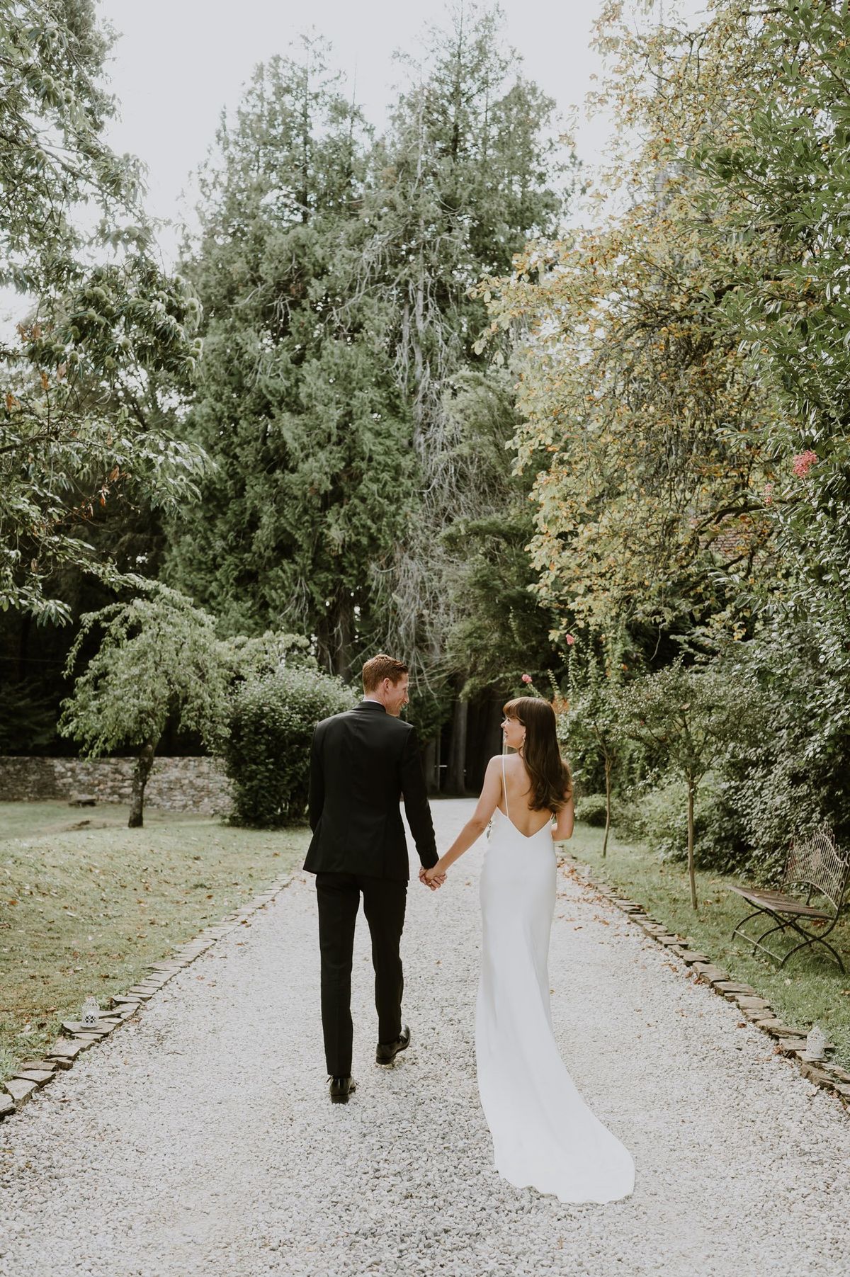 Couple walking hand-in-hand on gravel path bride in ivory slip gown looking back at groom in black suit