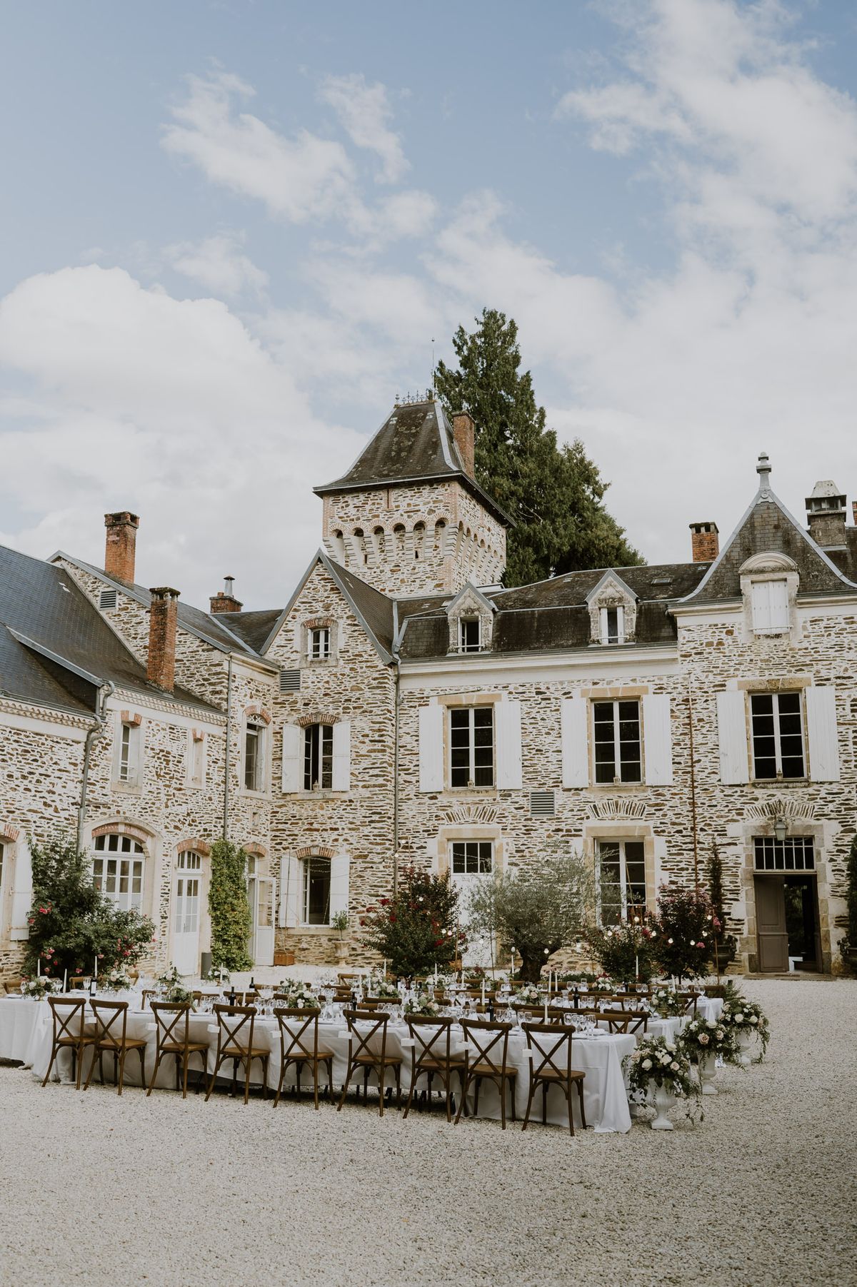 An outdoor wedding reception is set up in the gravel courtyard of a French château, with long banquet tables dressed in white linen and lined with dark-stained wooden cross-back chairs. The tablescapes include tall white taper candles, glassware, and low floral arrangements featuring blush, white, and soft pink blooms. Urn-style floral arrangements with blush and white flowers are placed at the ends of the tables, and potted olive trees and flowering shrubs are positioned between the tables and the building facade. The setting is a multi-wing French manor house built from rough-cut stone, featuring a central tower with decorative crenellation, slate-tiled mansard roofs, and arched window details. This is a wide establishing shot taken from ground level showing the full courtyard setup against the château facade. Potential venue feature image.