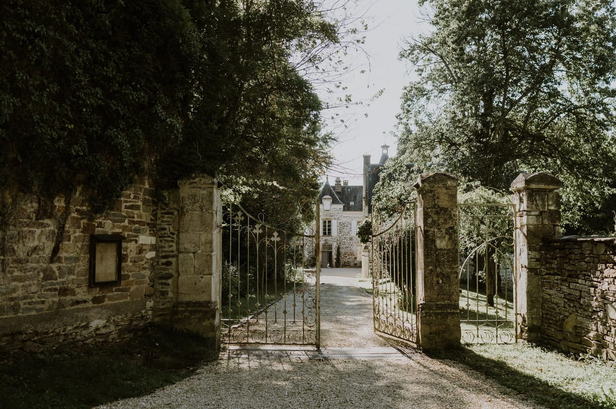 A wide shot looking through a set of open ornate wrought-iron gates mounted between aged stone pillars, leading down a gravel driveway toward a French manor or château in the background. The château building features steep slate rooflines, dormer windows, chimneys, and pale stone façade typical of historic French country architecture. No people are visible in the frame. The image has a warm, slightly muted tonal quality with dappled light filtering through the surrounding trees onto the gravel path. Potential venue feature image.