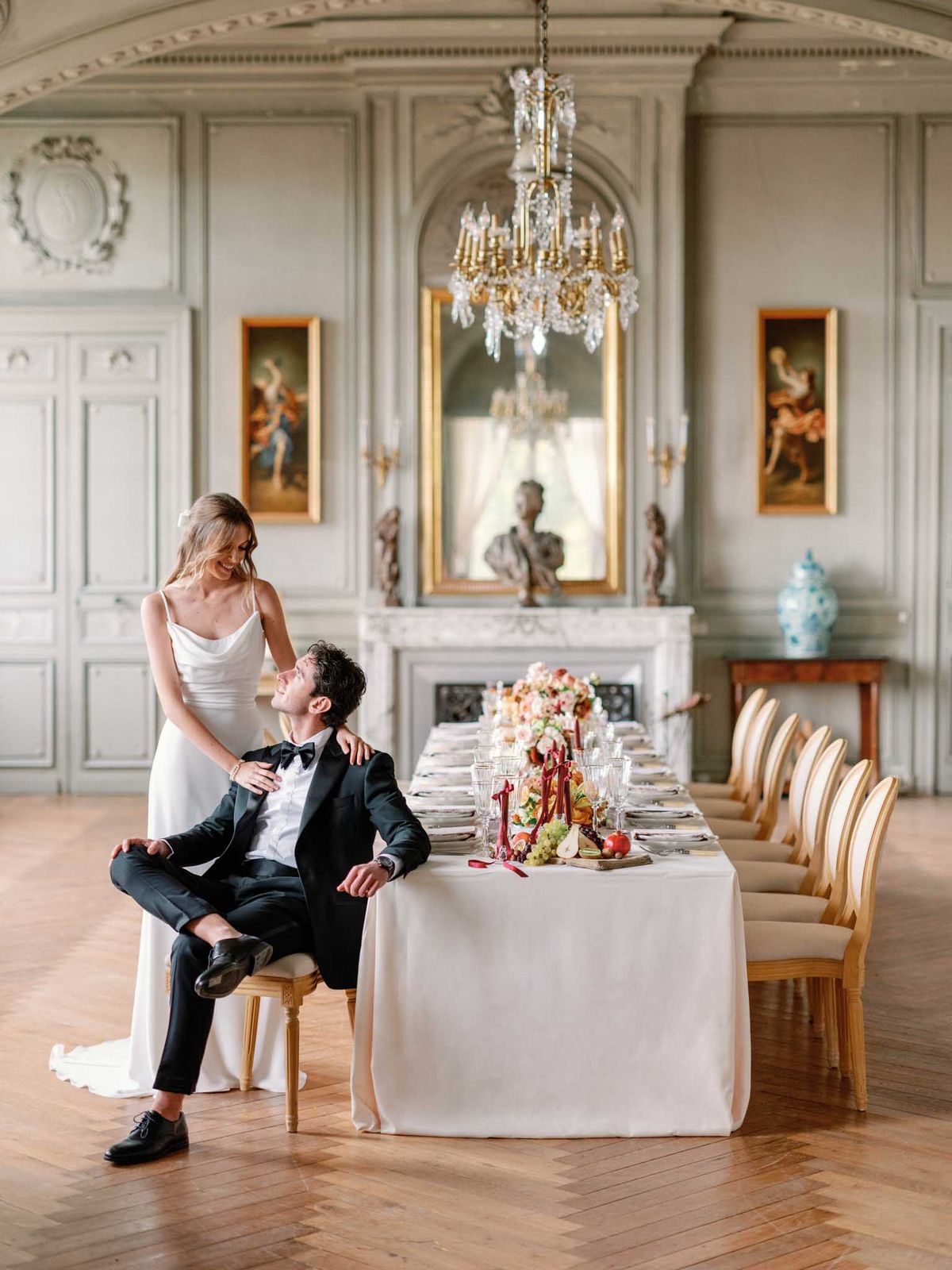 A bride and groom pose beside a long reception table inside a grand French château dining room with pale grey boiserie paneling, ornate plasterwork moldings, and a gold crystal chandelier overhead. The groom, wearing a black tuxedo with a black bow tie, sits casually on a gold Louis XVI-style chair while looking up at the bride, who stands behind him in a minimalist ivory cowl-neck slip gown with thin straps and a small white floral hair accessory. The long table is dressed in a white linen tablecloth and set with crystal glassware, white plates, and place settings, centered by a lush floral arrangement featuring coral, peach, and cream blooms alongside fresh fruits including grapes, pears, and apples, accented by deep crimson ribbon details and candles. The room features herringbone parquet flooring, gold-framed oil paintings in a classical style, wall sconces, a white marble fireplace with a sculptural bust and mirror above, and a blue-and-white ceramic vase on a side table, reflecting a classic French interior aesthetic. Wide portrait shot. Potential venue feature image.