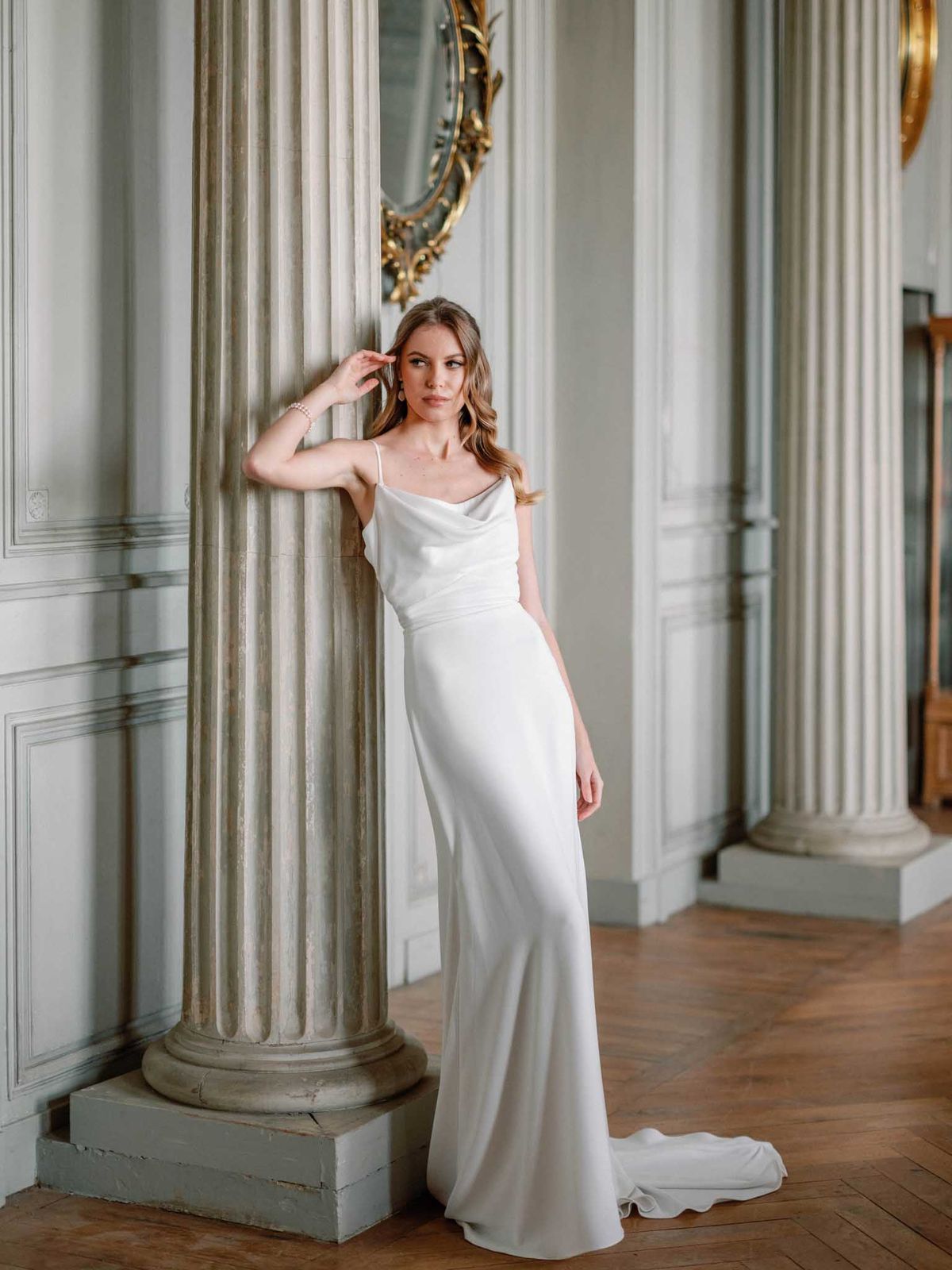 A bridal portrait of a single woman leaning against a fluted stone column inside a grand classical interior, likely a French château or hôtel particulier. She wears an ivory satin slip-style wedding dress with thin spaghetti straps, a cowl neckline, a fitted silhouette, and a short train pooling on the herringbone parquet floor. Her accessories include pearl drop earrings and a delicate pearl bracelet, and her hair is styled in loose waves. The background features pale grey-blue boiserie paneling, additional fluted columns, and a gilded ornate mirror, creating a classic French neoclassical aesthetic. The composition is a full-length portrait shot with soft, even natural light.