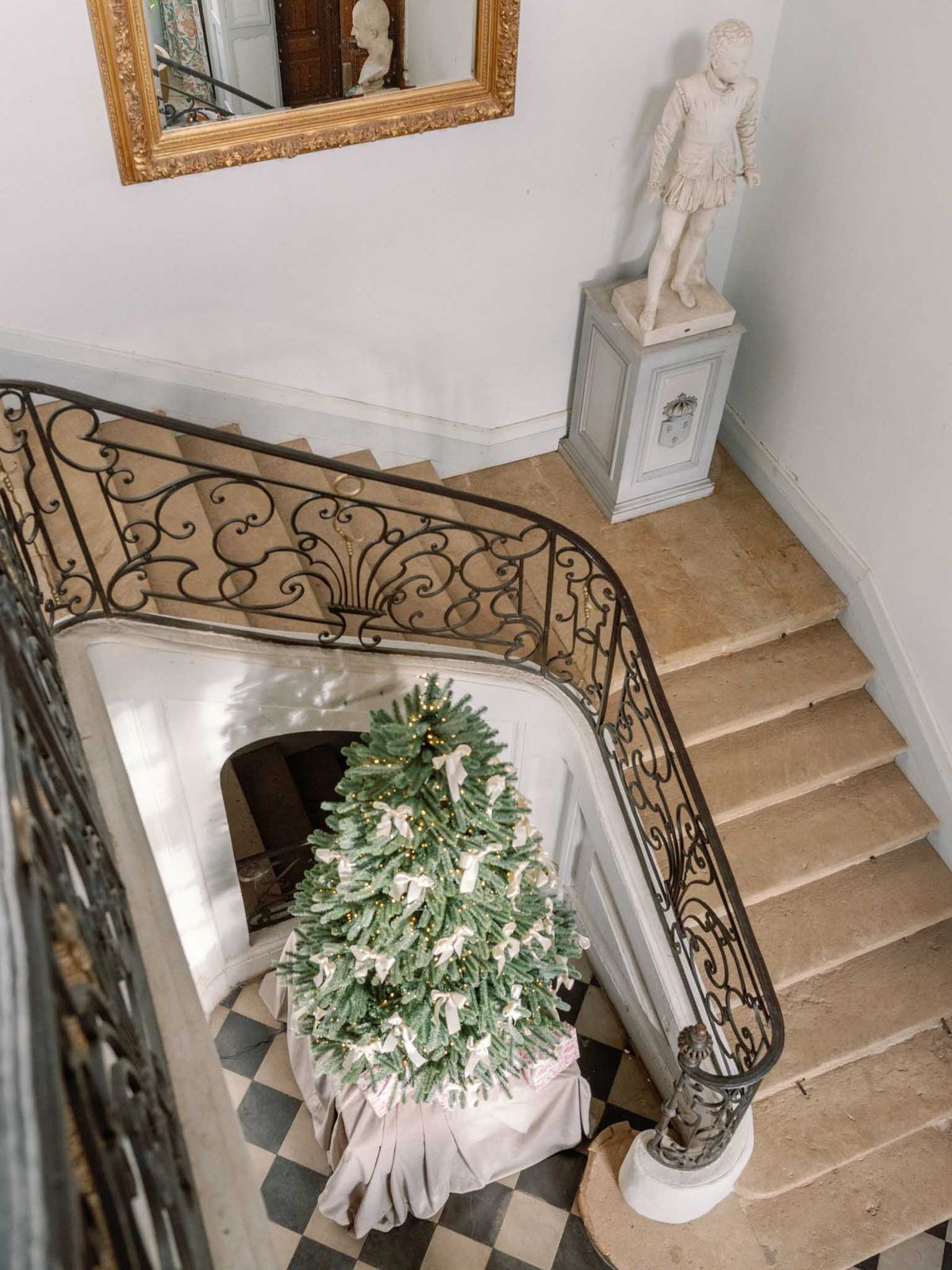 An overhead wide shot of a château interior staircase hall, captured from an upper floor looking down. The focal point is a large Christmas tree decorated with warm fairy lights and ivory ribbon bows, wrapped at the base with a linen-toned skirt, positioned at the foot of a curved stone staircase with an ornate wrought-iron balustrade featuring scrollwork detailing. The floor below is laid in a black-and-white diamond-pattern stone tile. A white marble statue of a classical figure stands on a pale grey carved pedestal on the landing. A large gold-framed mirror with carved detailing hangs on the white wall above. No people are present in the image. Potential venue feature image.