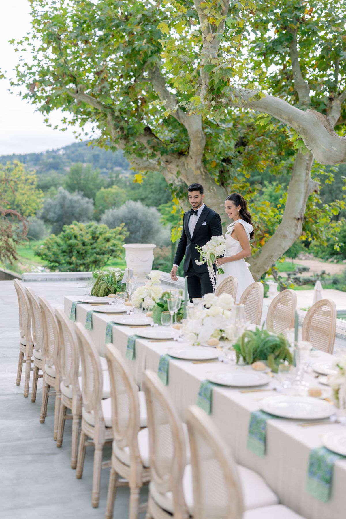 Bride and groom walking past outdoor reception table with cream linens, sage napkins, rattan chairs, and white floral centerpieces