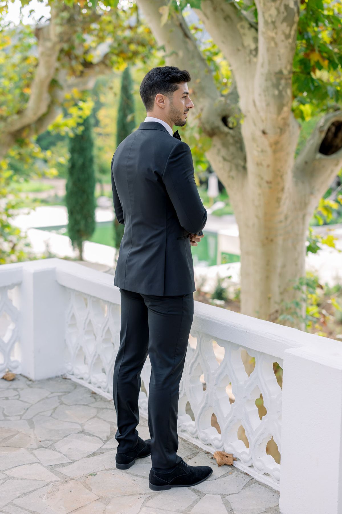 Groom in black double-breasted suit standing at stone balustrade terrace with cypress trees and garden path