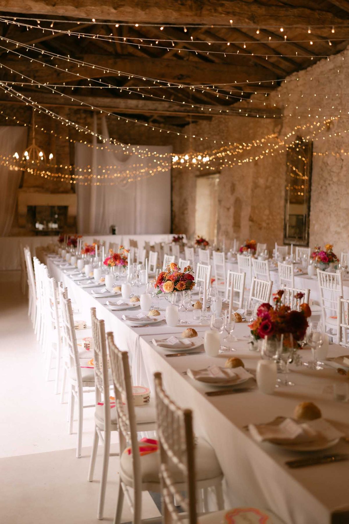 Rustic stone reception hall with fairy light ceiling, white banquet tables, and hot pink and orange floral centerpieces