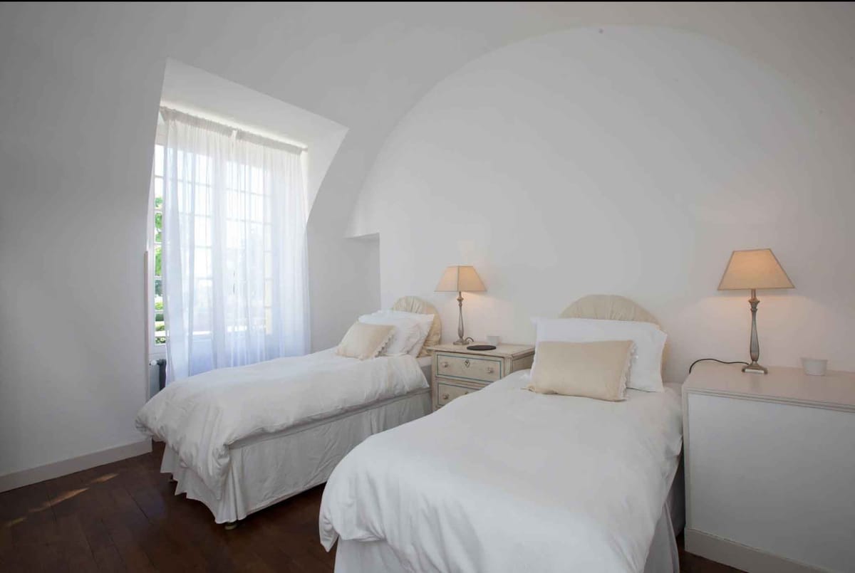Interior wide shot of a twin bedroom inside what appears to be a French château or manor house, featuring a vaulted barrel ceiling finished in white plaster. The room contains two single beds dressed in white linen with cream ruffled accent pillows and cream upholstered headboards, flanked by a painted grey-blue nightstand and a white bedside table, each topped with a warm-toned beige table lamp. A tall arched window with white sheer curtains allows natural daylight into the space, and dark hardwood flooring contrasts with the all-white walls and ceiling. The décor palette is white, cream, and soft grey with a classic French country styling. Potential venue feature image.