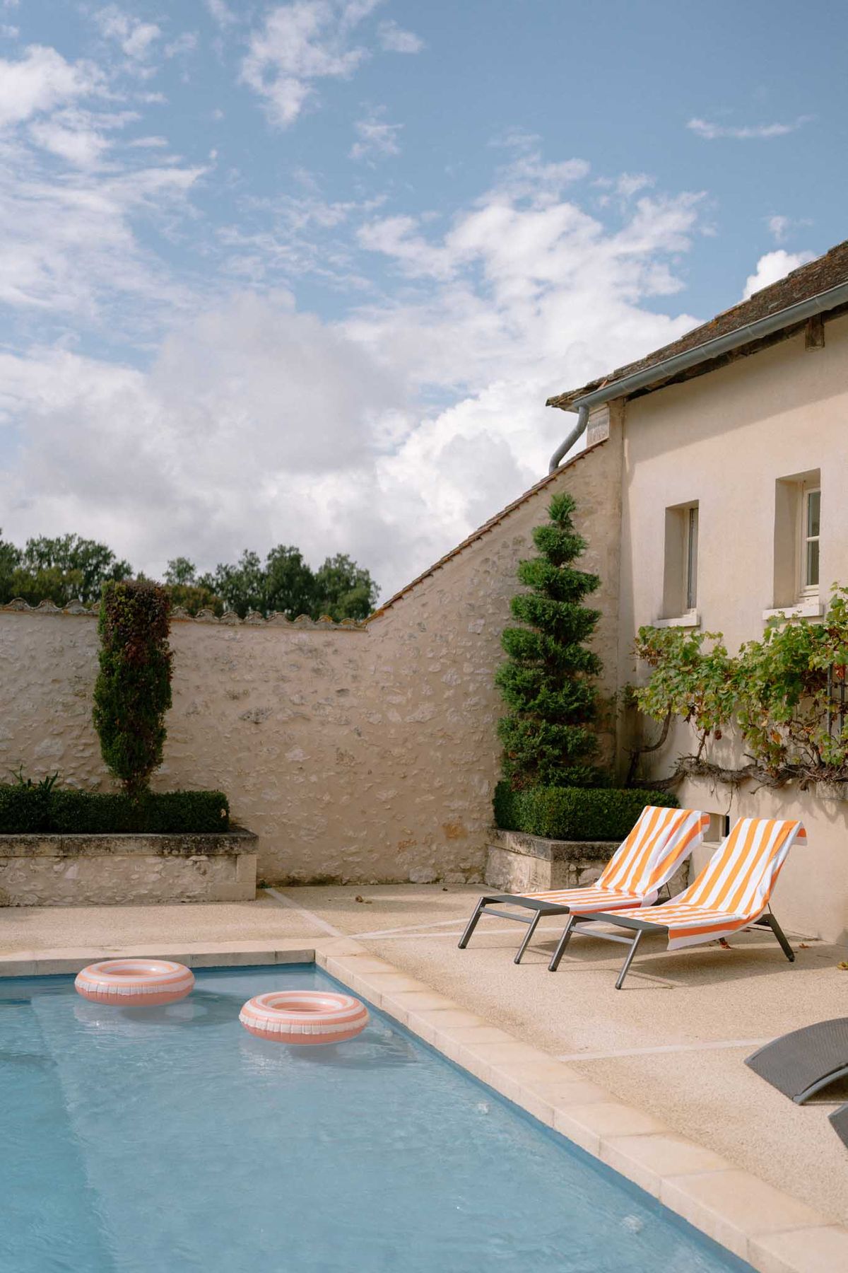 An outdoor pool area at a French country property, showing no wedding activity or people. Two sun loungers with orange and white striped towels are positioned on the poolside terrace, and two blush pink inflatable ring floats sit in the water. The pool is bordered by a pale stone surround, with clipped topiary spirals and boxwood hedging visible against a rendered stone boundary wall. A traditional French farmhouse or maison de maître occupies the right side of the frame, featuring a tiled roof and climbing vine. Wide shot composition. Potential venue feature image.