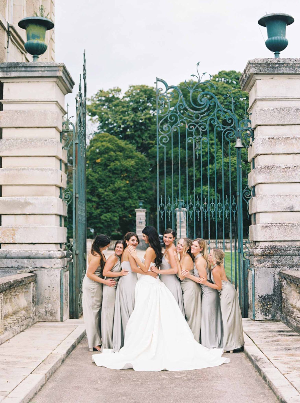 Bride and seven bridesmaids in taupe dresses posing symmetrically between stone pillars at ornate estate gate
