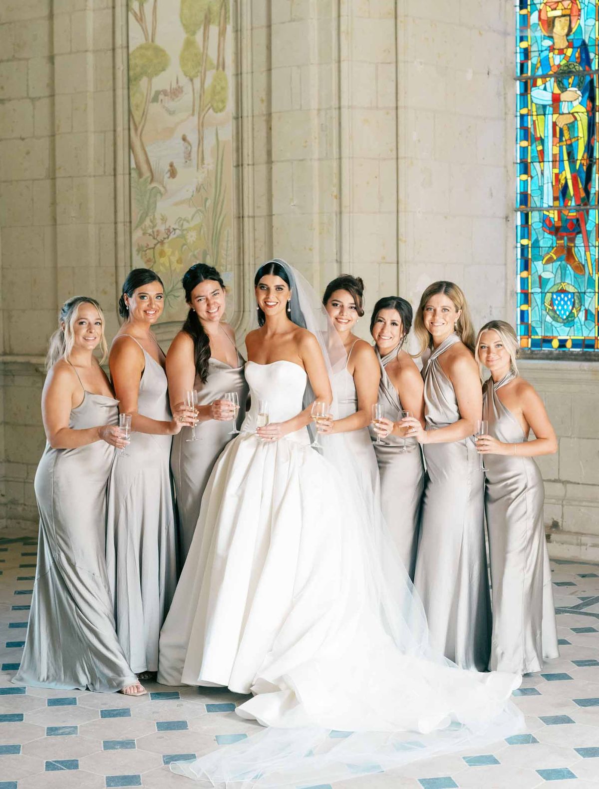 Bride in ivory ball gown with eight bridesmaids in taupe dresses holding champagne in a chapel with stained glass