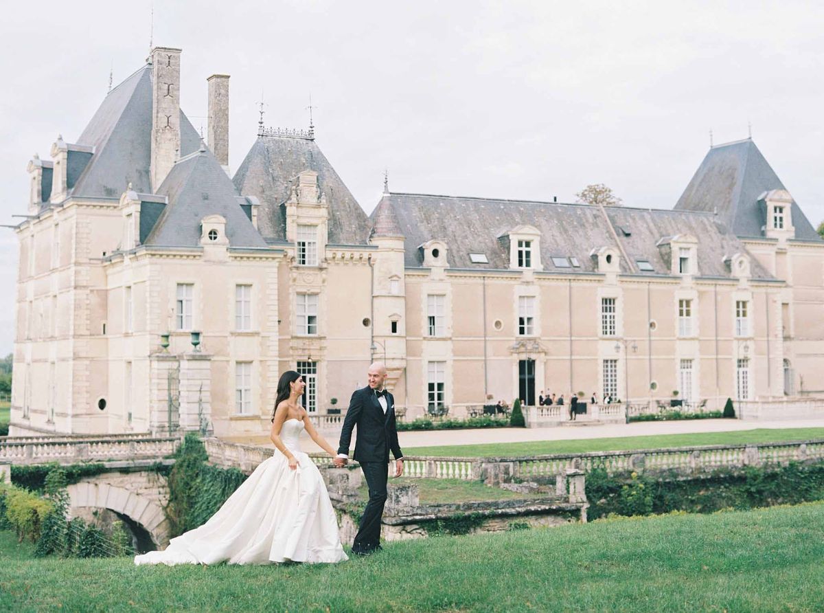 Bride in strapless ball gown and groom in black tuxedo on manicured grounds before limestone château with mansard roof